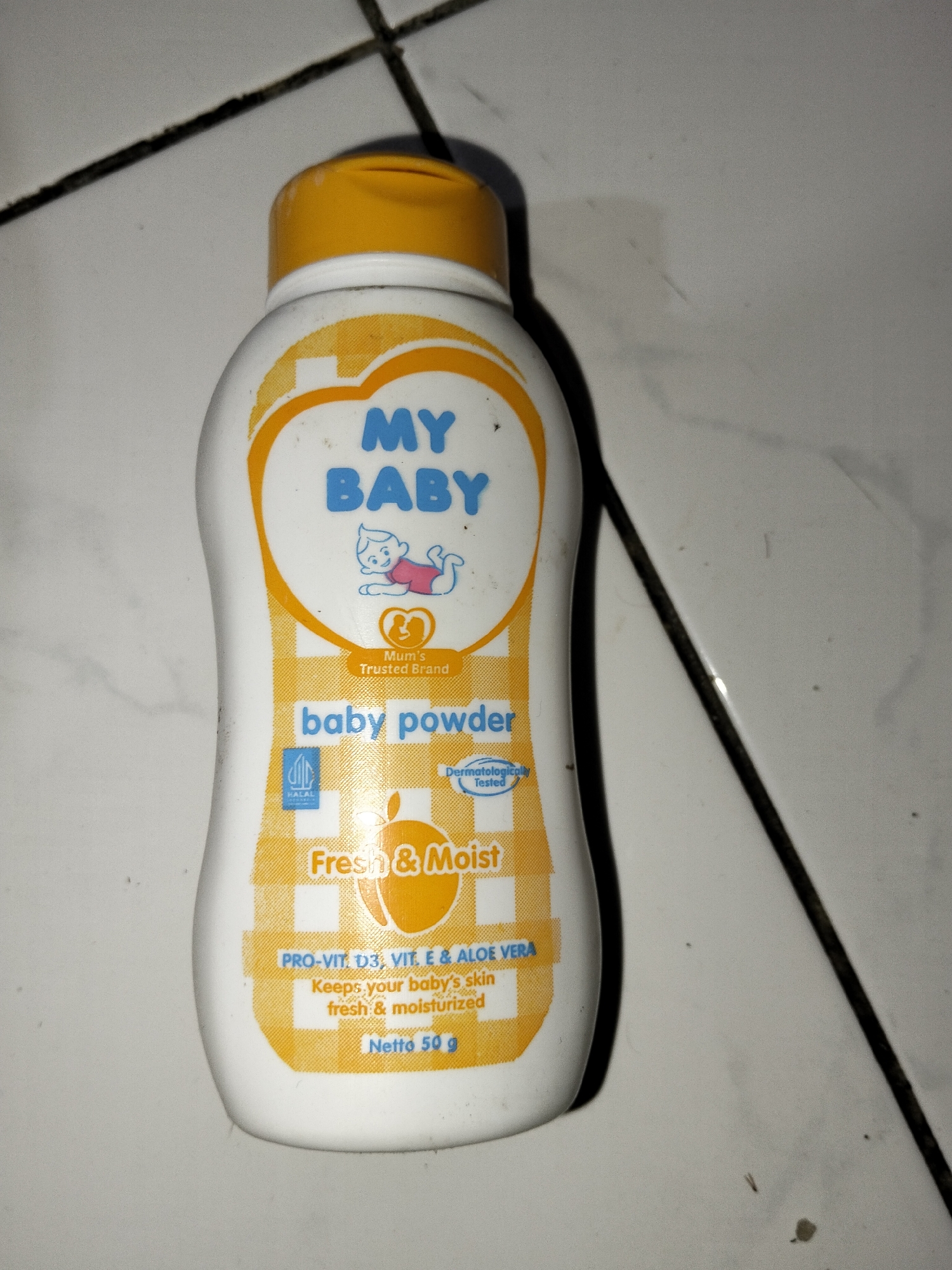 A white bottle of "MY BABY" brand baby powder is on a tiled floor. The bottle has a yellow cap and features blue text for the brand name. Below the brand name is an illustration of a baby. The label also states "baby powder," "Fresh & Moist," and lists ingredients like "PRO-VIT. D3, VIT. E & ALOE VERA." The net weight is indicated as "Netto 50 g." The tiled floor has dark grout lines. The image is undated and unlocated to a specific city or geographical region.