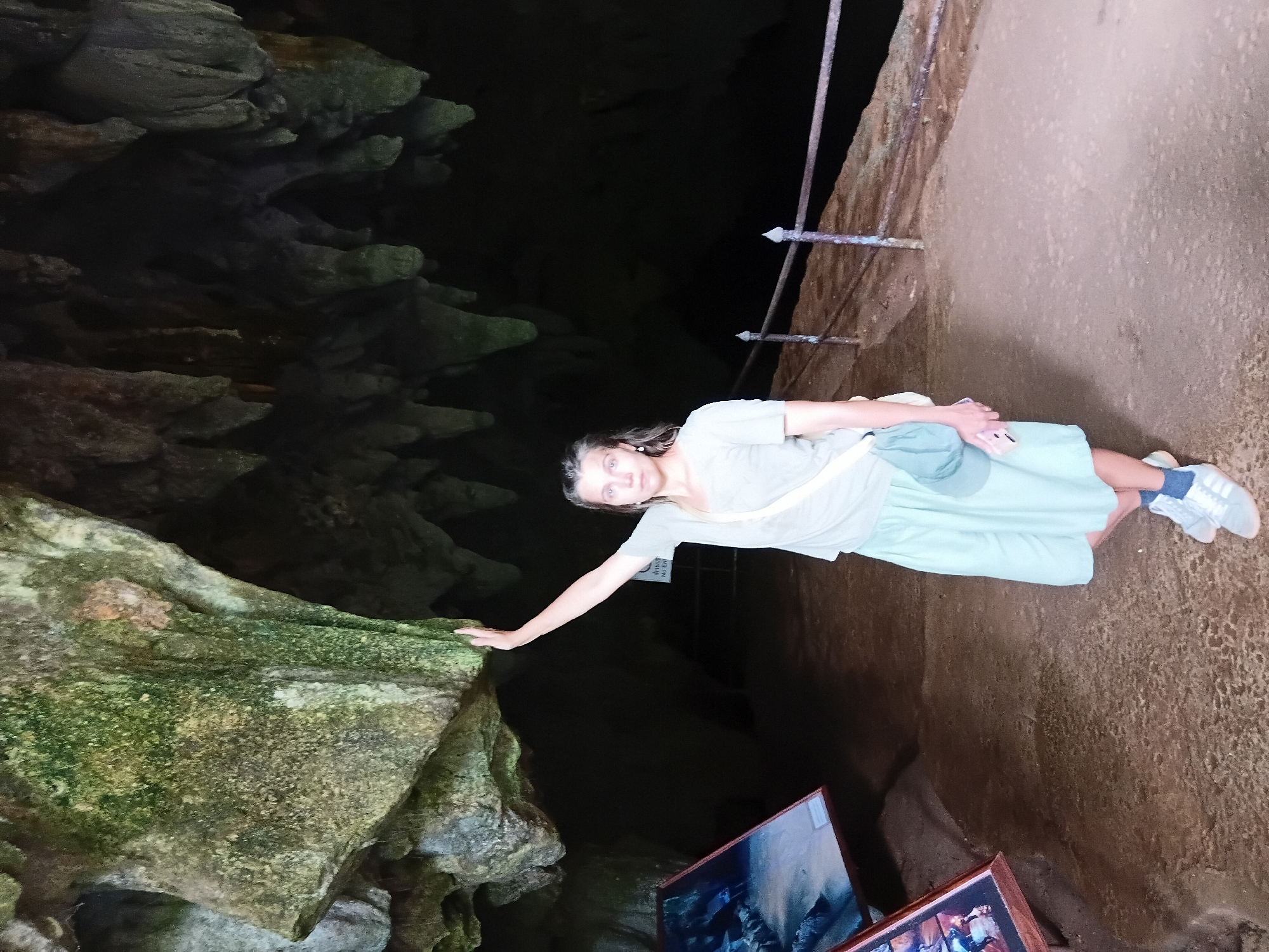A young woman with shoulder-length brown hair stands in a cave with her right arm extended upwards, touching the cave ceiling. She is wearing a light grey t-shirt, a light blue skirt, and grey sneakers with white laces. A light blue crossbody bag is slung over her right shoulder, and she is holding a pink smartphone in her left hand. The cave has rocky walls and a dark background. In the foreground, there is a pathway with a metal railing. To the left of the woman, there are two framed pictures displayed on easels. The scene appears to be within a cave in Mae Sai District, Thailand, likely a tourist attraction.