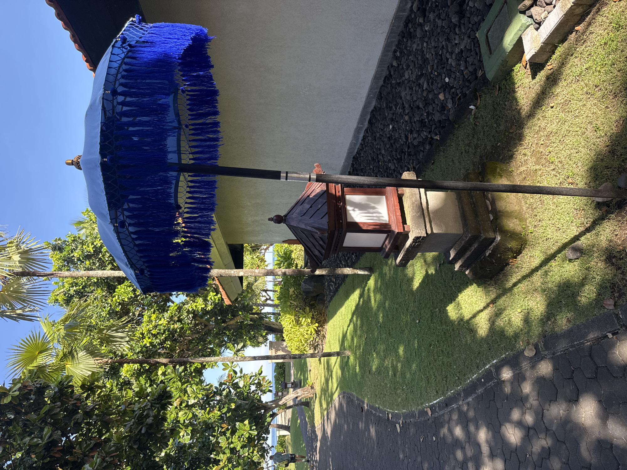 The image captures an outdoor scene in Kuta Selatan, Indonesia. A large blue fringed umbrella is prominently displayed, positioned next to a decorative outdoor lamp. The backdrop features a building with a light-colored wall bordered by dark gravel. Lush green grass and trees are visible, leading towards a distant view of the ocean. A paved pathway snakes through the landscape, with a person walking in the distance. The sky is clear and blue.