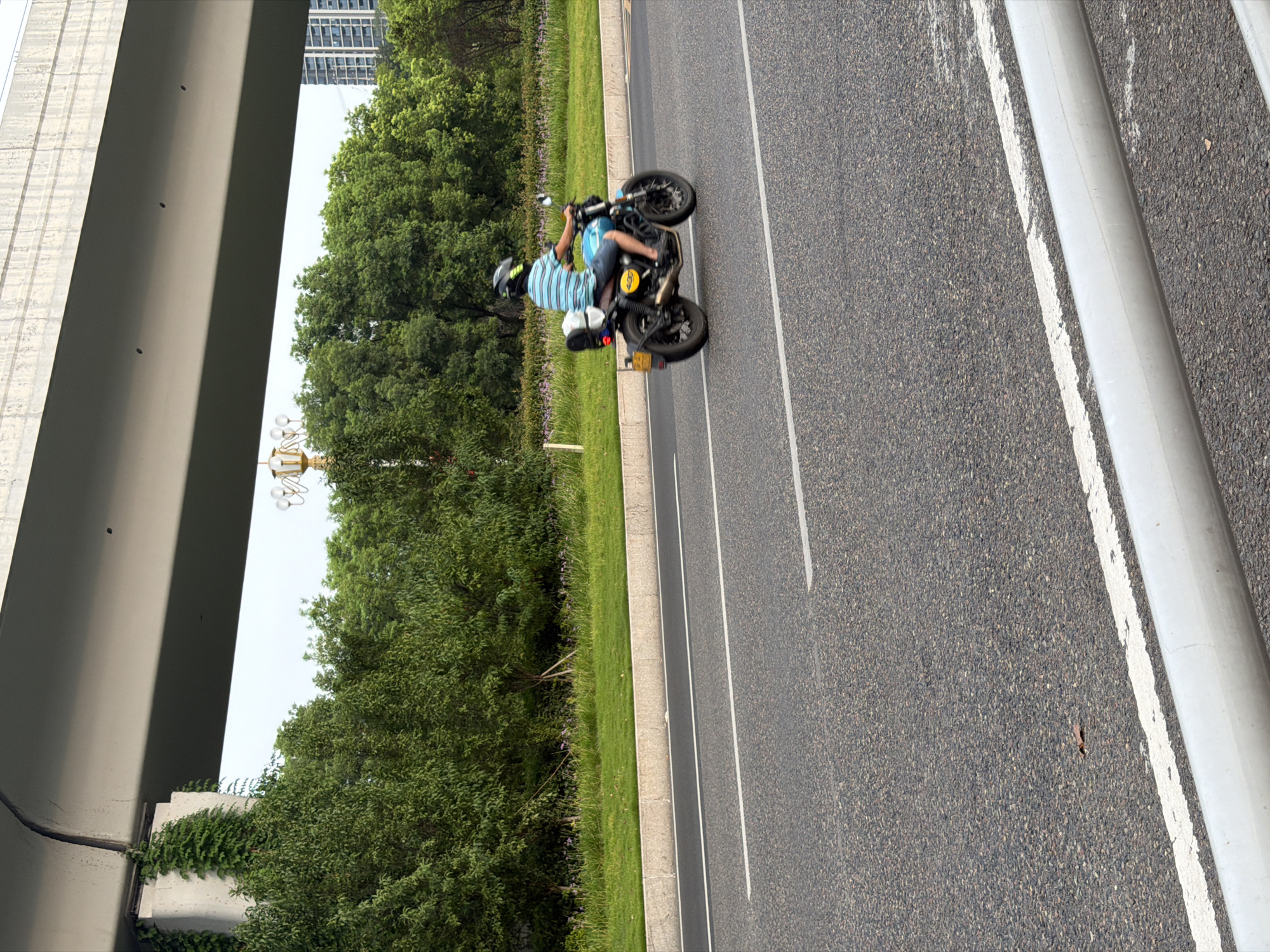 A person wearing a blue and white striped shirt, denim shorts, and a black helmet is riding a blue and black motorcycle on a paved road. The motorcycle has a yellow circular emblem with numbers on it and a white bag on the back. The road is bordered by a grassy median with trees and some small purple flowers. In the background, there is a large elevated concrete structure, likely an overpass, and beyond that, several tall buildings. The scene appears to be in an urban or suburban area of Jiangjin, China.
