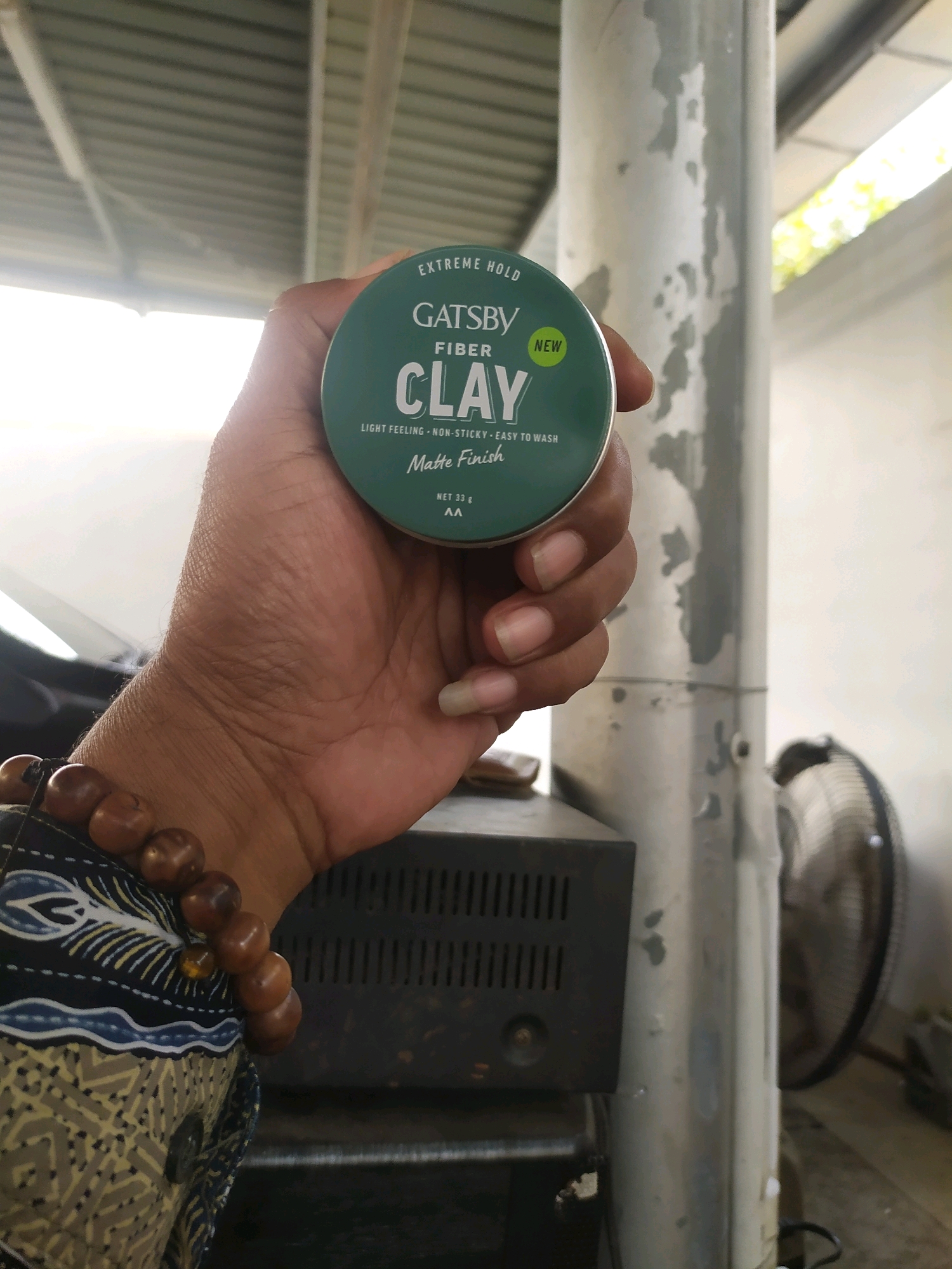 The image depicts a hand holding a Gatsby Fiber Clay hair product. The product is in a green circular container with white and green text. The hand is adorned with a beaded bracelet. In the background, a metal pole and a fan are visible. The scene appears to be outdoors, possibly under a roofed structure, with a bright sky in the upper portion of the image. The overall setting suggests a casual, perhaps residential or workshop environment in Sleman, Indonesia.