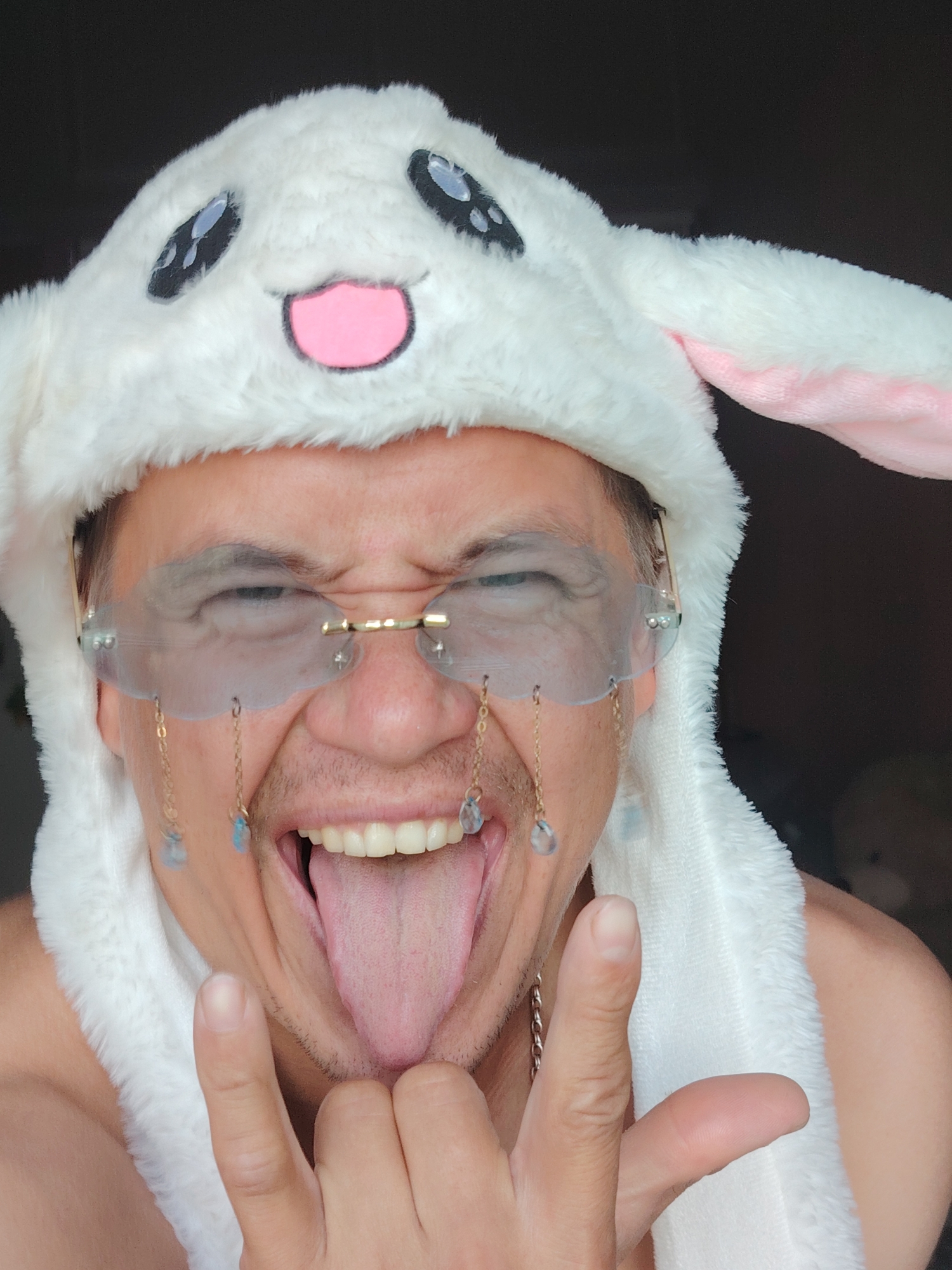A person is wearing a white plush bunny hat with large embroidered eyes and a pink nose. They are also wearing clear, rimless glasses with gold accents, from which hang two delicate chains adorned with small blue crystals. The person's mouth is open, with their tongue sticking out, and they are making a rock-and-roll hand gesture with their right hand. The background is dark, with a hint of a soft toy on the right side. There is no indication of a specific location within Chaikovsky, Russia.