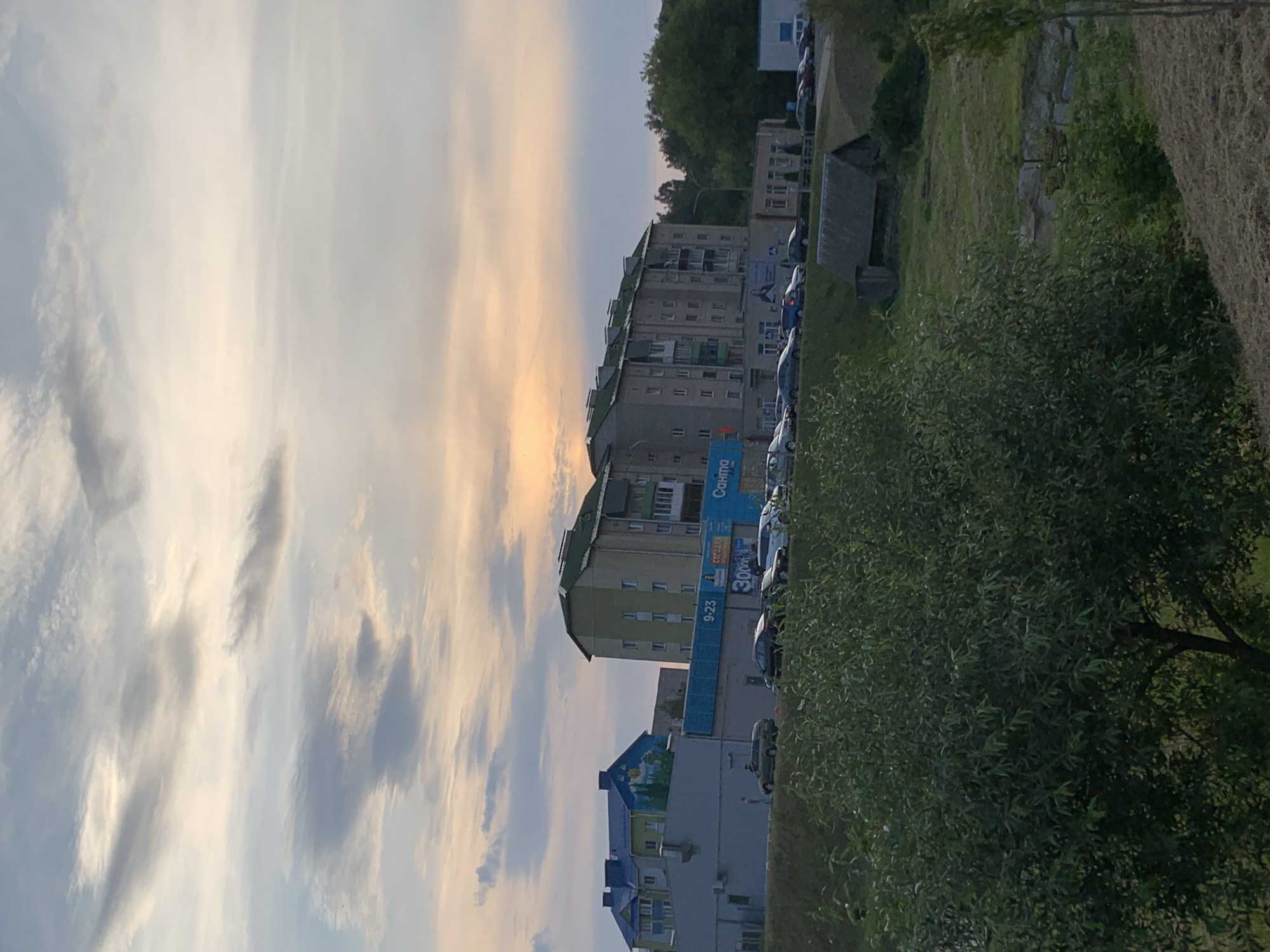 This is a medium-shot, eye-level photograph taken outdoors at dusk in the city of Kapyĺ, Belarus. The sky is a gradient of pale yellow and pink, with scattered grey clouds. In the foreground, there is a large, leafy green tree on the left side of the image, partially obscuring a sloped embankment. Behind the tree, several multi-story apartment buildings with various colored roofs are visible. The buildings are predominantly grey and light green, with some featuring blue accents. A blue banner with white text and advertisements is present on one of the buildings. A parking lot with several cars is situated in front of the buildings. Further in the background, a small, weathered wooden structure with a dark roof sits on a grassy hill. The overall scene depicts a residential area as daylight fades.