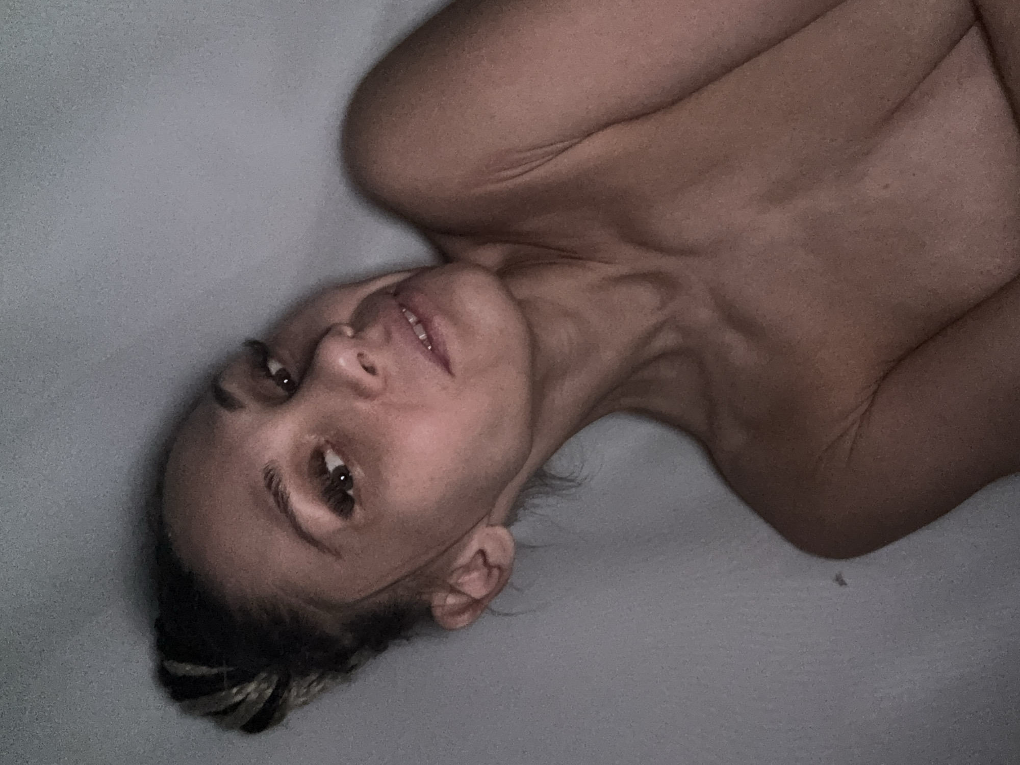 A woman is lying on a gray surface. Her head is tilted to the side, and she is looking at the camera. Her hair is braided, and she is wearing little clothing. The location is not identifiable from the image.