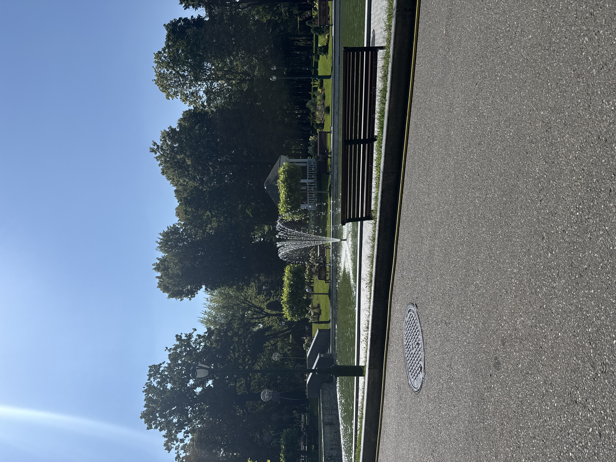 A sunny day in a park with a fountain and a gazebo. In the foreground, there is a paved path with a manhole cover and a wooden bench. A pond with a fountain is visible in the middle ground, surrounded by manicured lawns, trees, and more benches. In the background, there are tall trees and a white gazebo. The sky is clear and blue with a bright sunbeam. The overall scene depicts a peaceful, urban park setting.