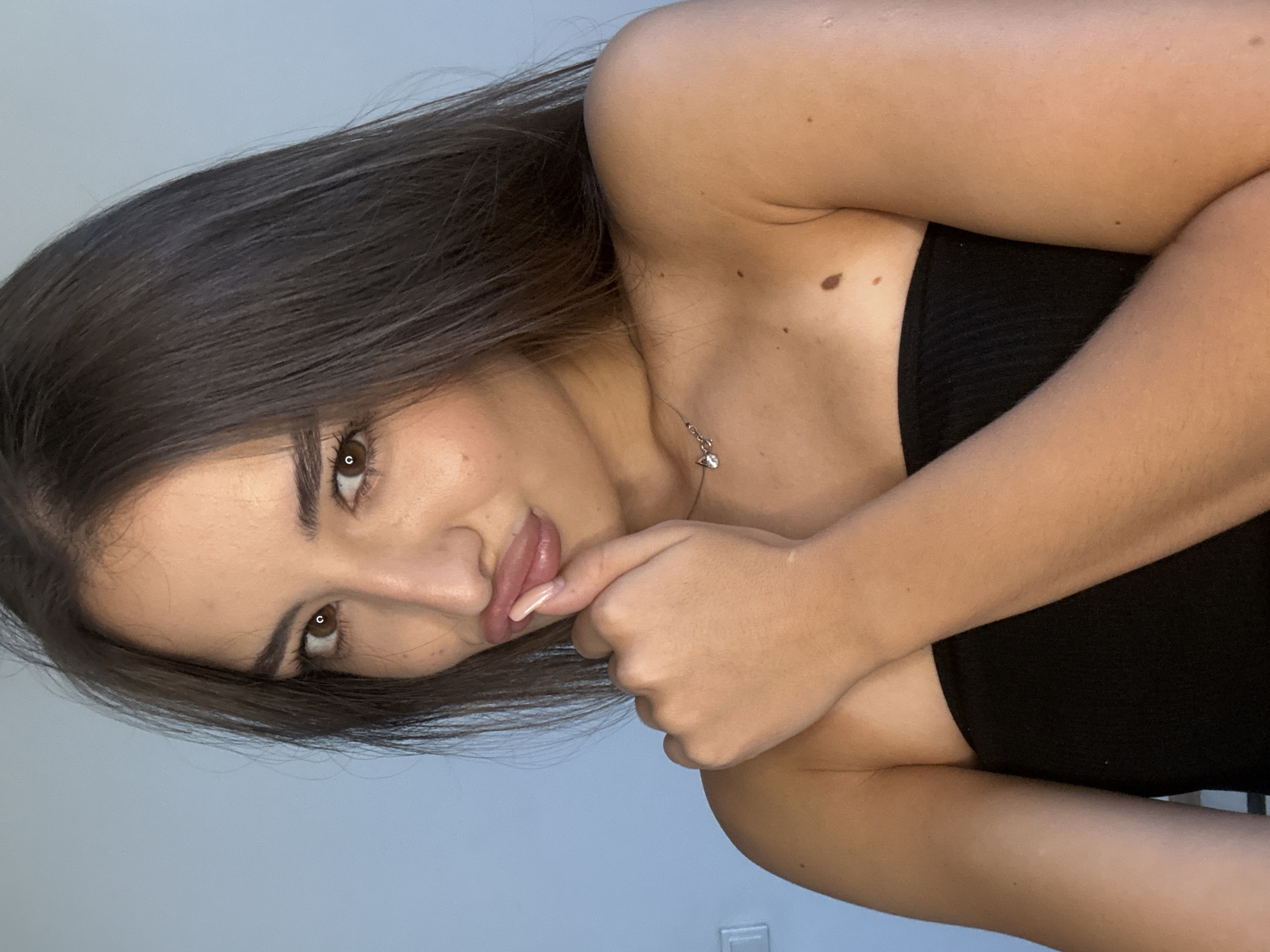 The image is a medium shot of a young woman with long, dark brown hair. She is looking directly at the camera with brown eyes and her lips are slightly parted, with her right index finger touching them. She is wearing a black strapless top. A small, silver necklace with a charm is visible around her neck. The background is a plain, light blue wall. There is a light switch on the wall to the left of the woman. The woman has several moles on her right shoulder and upper arm. The image was taken indoors in what appears to be a residential setting. There is no indication of the location being in Chișinău, Moldova.