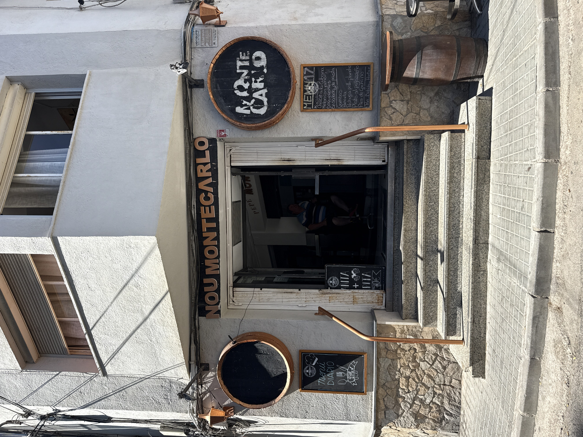 This is a vertical outdoor photograph of the exterior of a restaurant named "Nou Montecarlo" in Blanes, Spain. The building has a white stucco facade and a row of stone steps leading up to the entrance. The entrance is a wide doorway with metal shutters partially pulled down. Above the entrance, there is a round sign shaped like a barrel with "MONTE CARLO" written on it. To the right of the barrel sign, a chalk menu board lists daily specials.
To the left of the entrance, a vertical sign reads "NOU MONTECARLO" in large, dark letters with lighter outlines. Below this sign, another barrel-shaped object, possibly a planter or decoration, is visible. The steps are made of speckled grey stone, and a stone wall with irregular shapes lines the side of the staircase. A wooden barrel rests on the stone wall to the right of the entrance, serving as a decorative element.
Several smaller chalkboards are placed near the entrance, one showing "MENÚ 17" with a list of food items, and another with "ANITA + PITA 70€". Another chalkboard to the left of the entrance displays "MENÚ DIARIO" with icons of food and drink. The lighting suggests it is daytime, likely sunny, casting shadows on the building and steps. Electrical wires are visible near the ground level on the left side of the image, indicating the urban setting. The overall impression is that of a casual, possibly traditional, eatery. No people are clearly visible in the main shot, though a person can be glimpsed inside the dimly lit restaurant.