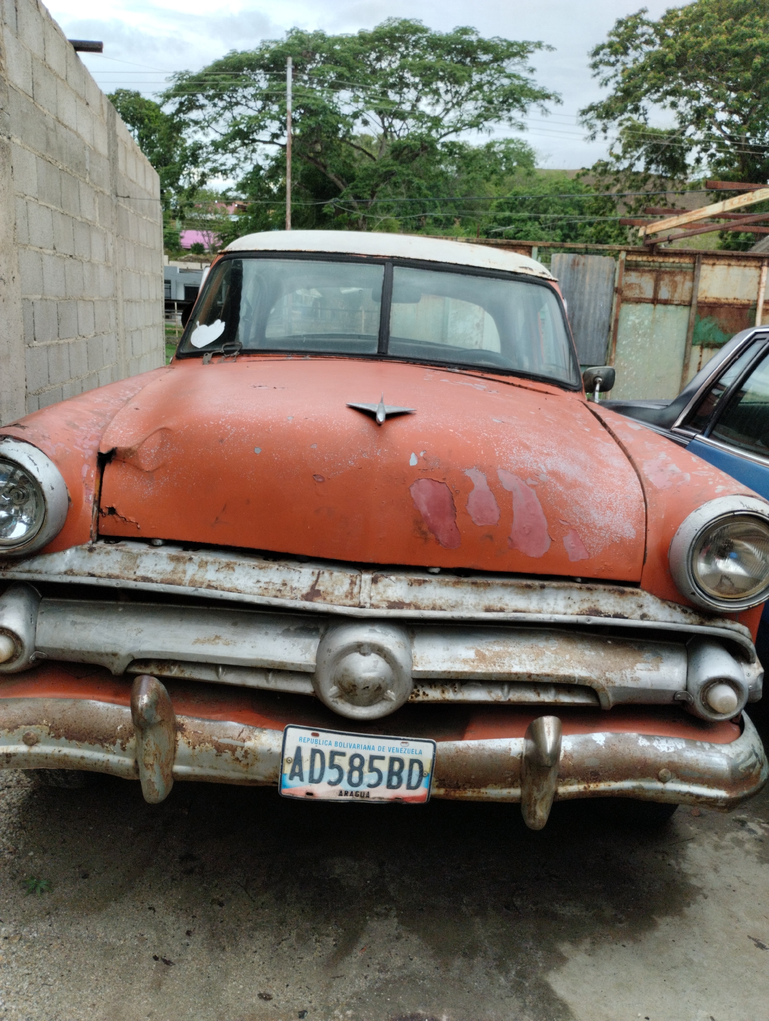 The image captures a front view of an old, heavily weathered car, likely a classic American model, parked outdoors. The car is predominantly a faded, peeling orange-red with a white roof. Its paint is extensively chipped, revealing lighter primer spots, and there are visible dents and rust on the hood, grille, and bumper. The chrome grille and bumper are also heavily rusted and corroded. Large, circular headlights are on either side of the front. A small, stylized hood ornament is present in the center of the hood.
To the left of the car is a concrete block wall, suggesting an enclosed area or a building. In the background, beyond the car, are lush green trees and an overcast sky, indicating daytime with diffused light. To the right, another vehicle, partially visible and dark blue, is parked next to the old car, along with a rusted corrugated metal fence or structure.
A Venezuelan license plate is affixed to the front bumper. The visible text on the license plate reads:
"REPUBLICA BOLIVARIANA DE VENEZUELA"
"AD585BD"
"ARAGUA"
The scene depicts a static moment, with the car appearing to be disused or in a state of disrepair. The overall impression is one of age and neglect, set in an outdoor, somewhat rustic environment in La Aguadita, Venezuela.