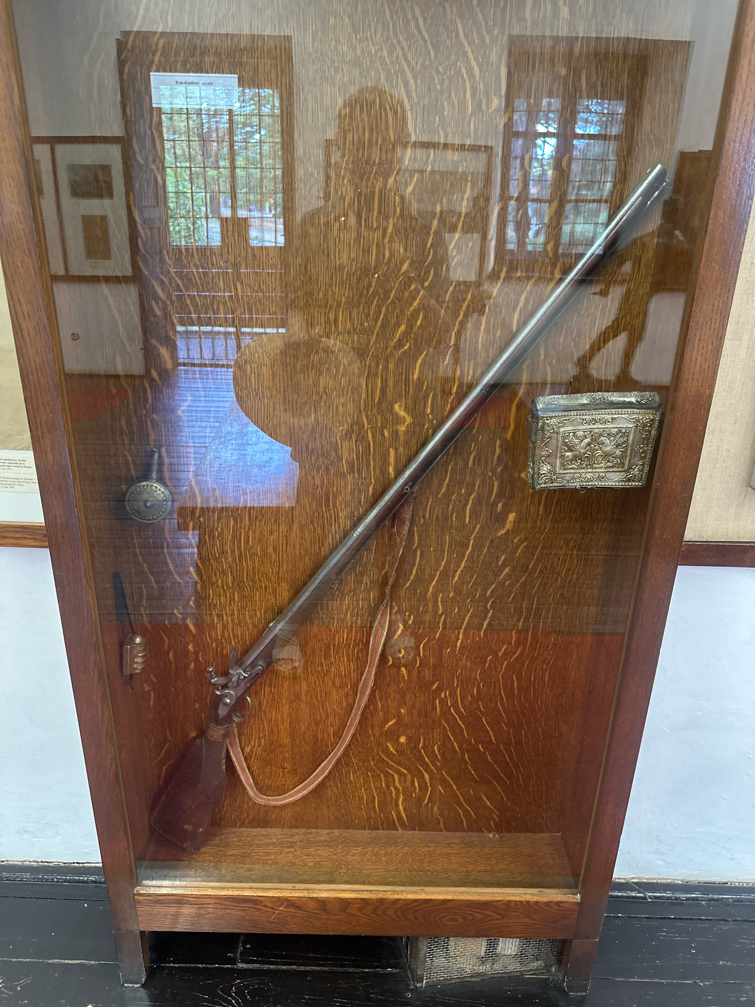 The image displays a glass-fronted wooden display case containing a vintage firearm. The case is made of dark wood with a visible grain pattern. Inside, a long rifle with a dark wooden stock and metal barrel is prominently featured, positioned diagonally from the lower left to the upper right. A light-colored strap is attached to the firearm. To the left of the rifle, a small, ornate metal object, possibly a flask or powder horn, is suspended. Further down on the left, a small, dark object with a ribbed texture is also attached. To the right of the rifle, a small, intricately decorated metal box with a raised relief design sits on the wooden backing of the case. The background of the display case shows reflections of a window with multiple panes, suggesting an outdoor scene with greenery outside. There are also faint reflections of human figures, indicating the presence of observers. On the far left of the image, a portion of a framed document or picture is visible. The display case is situated in what appears to be an indoor setting, possibly a museum or historical exhibit, with a light blue wall visible behind it and a dark wooden floor at the bottom. The context of Topola Municipality, Serbia, suggests this might be a historical artifact displayed within a local museum or cultural institution.