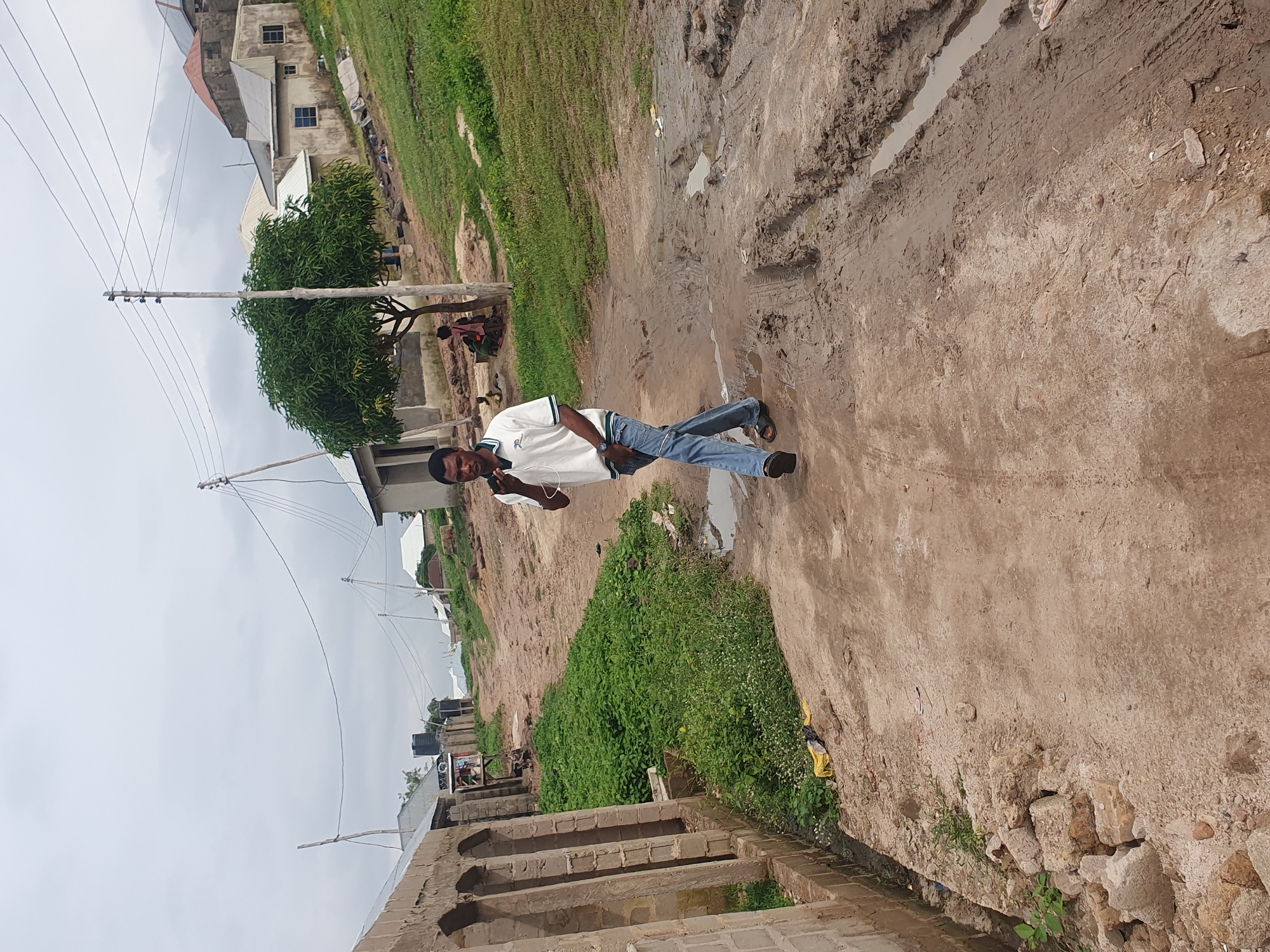 A man is walking down a dirt road in Lokoja, Nigeria. The road is muddy and rutted, with puddles of water. There are buildings on either side of the road, some of which appear to be unfinished. There is also a tree in the middle of the road. The sky is overcast.