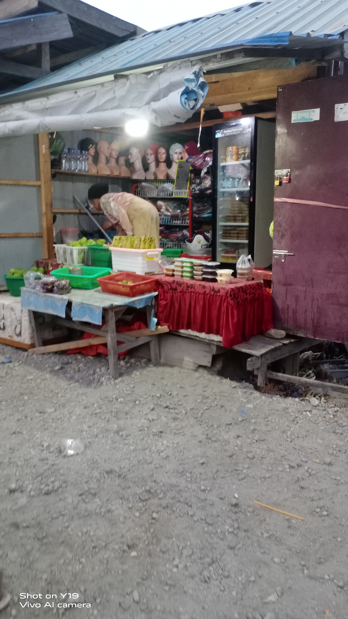 The image depicts an outdoor market stall, likely located in the city of Jati, Indonesia. The stall is situated on a gravel surface. In the foreground, a table draped with a red tablecloth displays various food items in small containers. Behind this table, another wooden table holds baskets of fruits and some packaged goods. A person, partially visible, is seen arranging items at the stall. To the right, a large, dark-colored refrigerator is prominent, with shelves stocked with items. The background shows a roofed structure extending over the stall, with mannequins displaying headwear visible on a shelf. The overall scene suggests a small, informal retail environment.