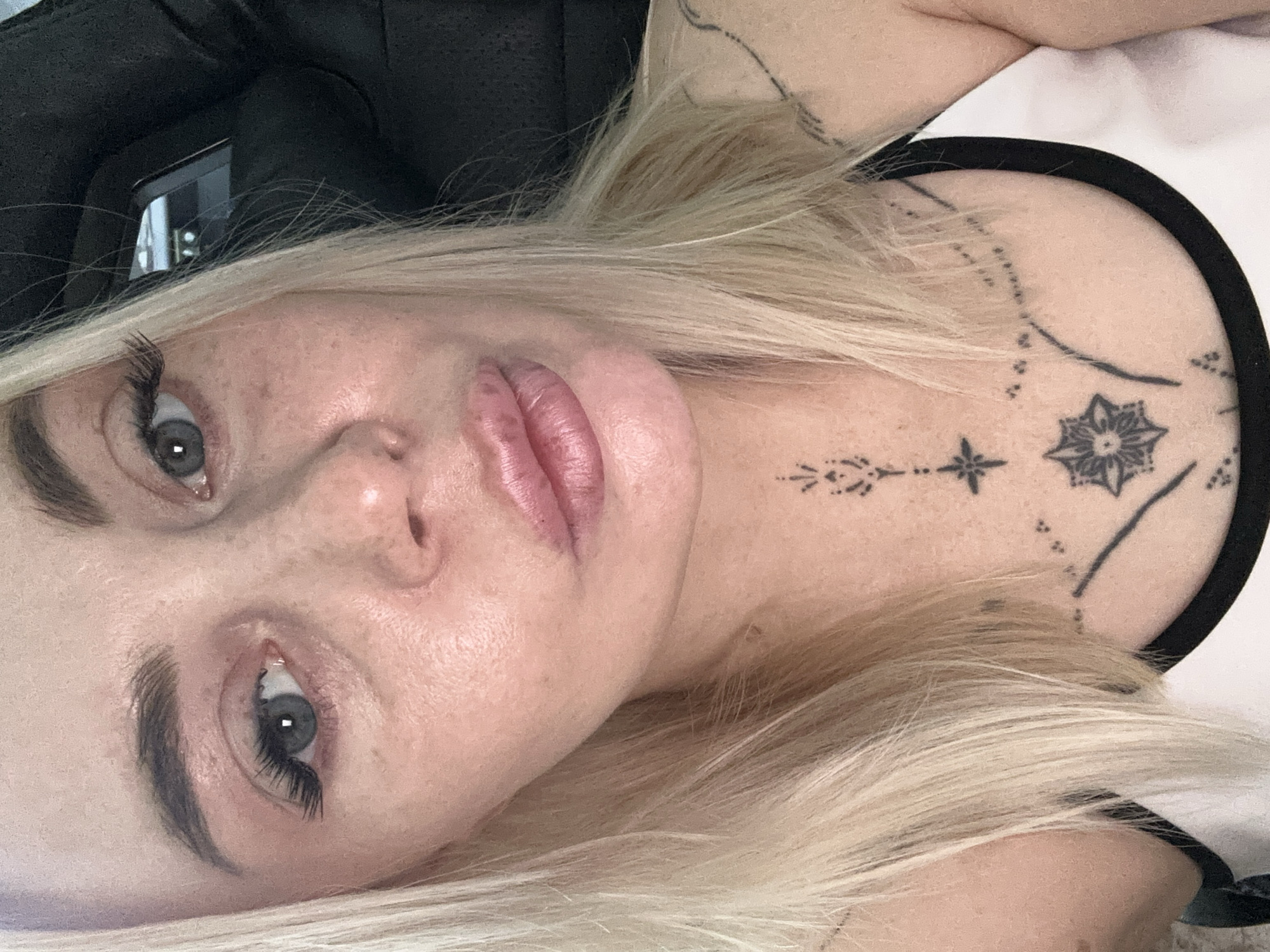 A close-up, head-and-shoulders shot shows a fair-skinned woman with blonde hair and blue-grey eyes. Her hair is parted on the left and falls to her shoulders. She is wearing makeup, including false eyelashes and lip gloss. She has freckles across her nose and cheeks. A dark tattoo is visible on her neck and chest, with smaller tattoos extending onto her shoulders. The background is dark and out of focus, with a hint of a black chair with a headrest on the right side. The woman is looking directly at the camera with her lips slightly parted.