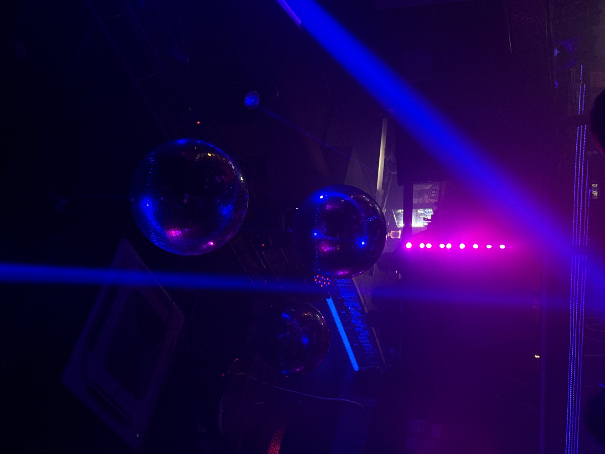 The image depicts the interior of a dark entertainment venue located in Perm, Russia. Dominant artificial lighting projects beams of vivid blue and magenta/purple. Three large, spherical disco balls hang from the ceiling, reflecting these colored lights. A rectangular, ceiling-mounted air conditioning unit is visible in the upper left. Multiple blue laser-like light beams traverse the scene, including two vertical beams in the upper and middle areas and one diagonal beam originating from the lower right. A vertical fixture containing eight illuminated magenta-pink lights is centrally positioned in the lower half. A horizontal blue light strip illuminates a background area consistent with a bar or stage, where bottles are faintly discernible. A Cyrillic sign reading "ВЫХОД" (EXIT) is visible in the lower left. Silhouetted individuals are present in the lower foreground, with one person's hand visibly raised on the left side, indicating activity within the establishment.