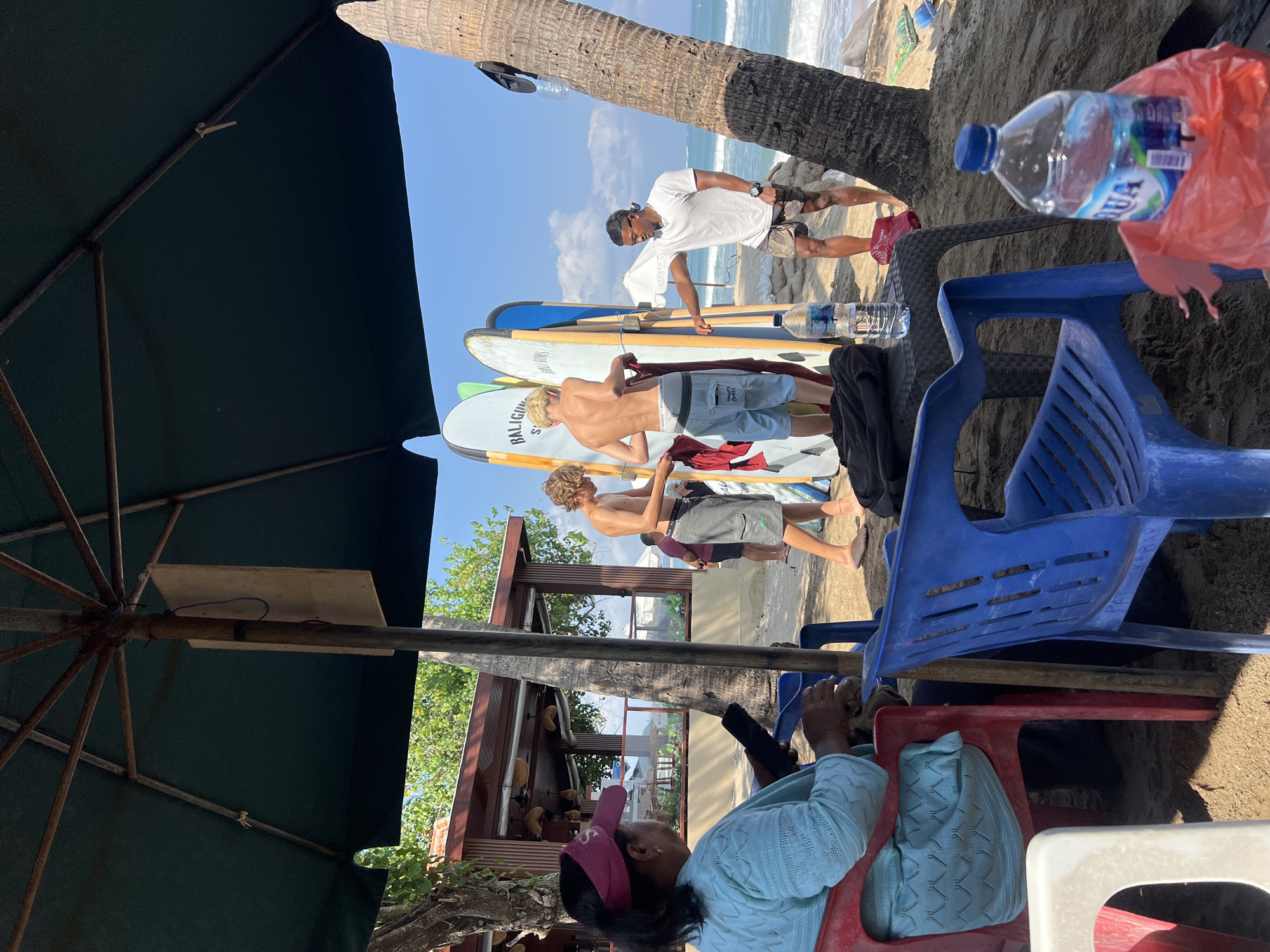 The image depicts a beach scene in Kuta, Indonesia, likely near a surf school or rental shop. In the foreground, under a large umbrella, a woman with dark hair and a pink visor sits in a red chair, looking at her phone. Next to her, a blue plastic chair is occupied by unseen individuals. A table holds a bottle of water. Behind them, two young men, presumably surfers, stand near several surfboards. One is changing out of his shorts, while the other appears to be assisting him. Further back, a man in a white t-shirt and shorts, possibly an instructor, gestures towards the surfboards. The background shows the ocean with waves, a sandy beach, and palm trees, with a building that resembles a restaurant or shop. The sky is clear and blue.