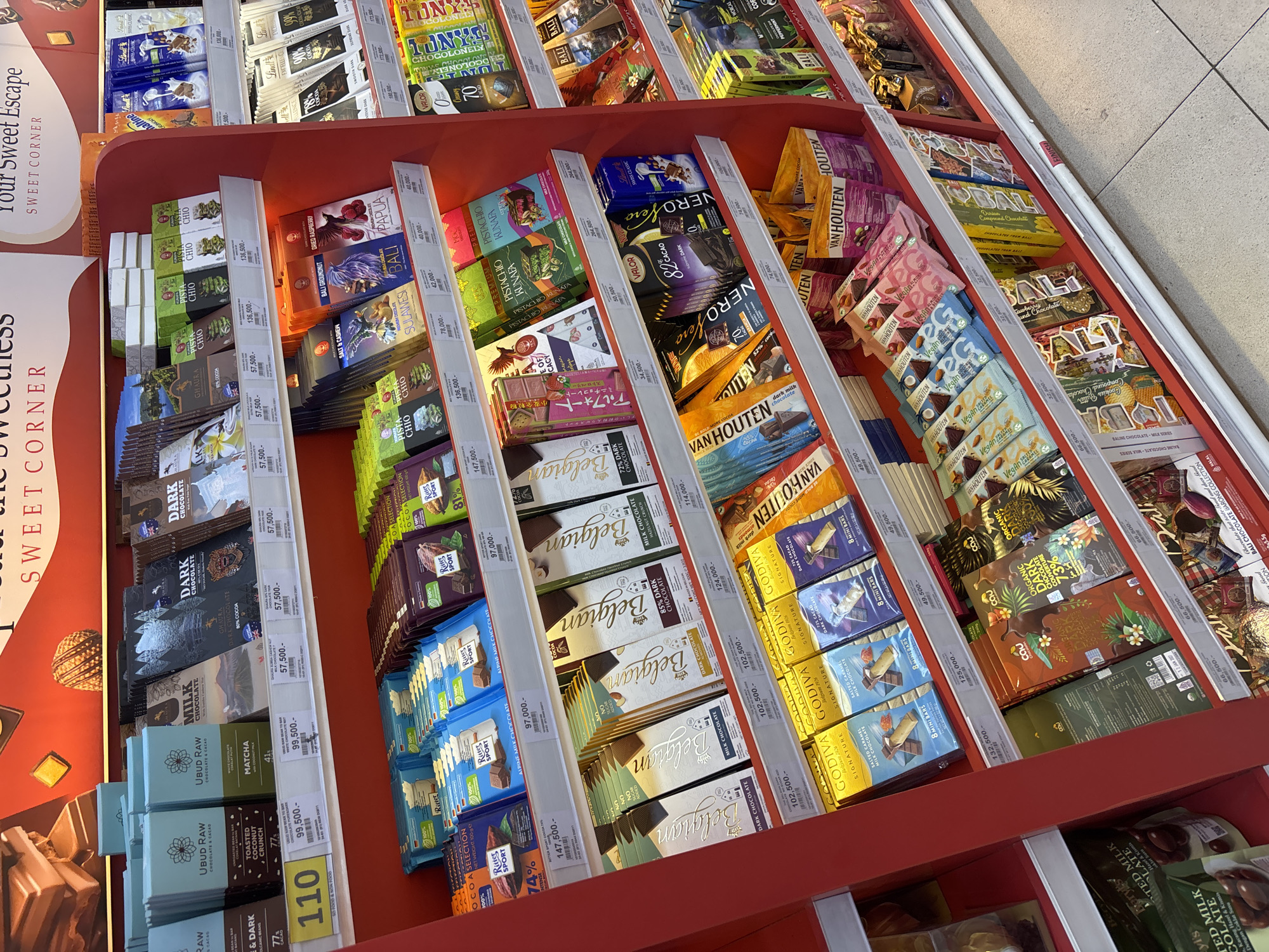 The image shows a section of a retail display, likely in a store located in the Kuta Selatan region of Bali, Indonesia, filled with various chocolate bars. There are multiple brands and varieties of chocolate, arranged in rows with visible pricing labels. Brands like UBUD RAW, Ritter Sport, Belgian, Van Houten, Godiva, and Tony's Chocolonely are displayed. Price tags are visible. The display itself is a red multi-tiered shelf. The floor appears to be light-colored tile.