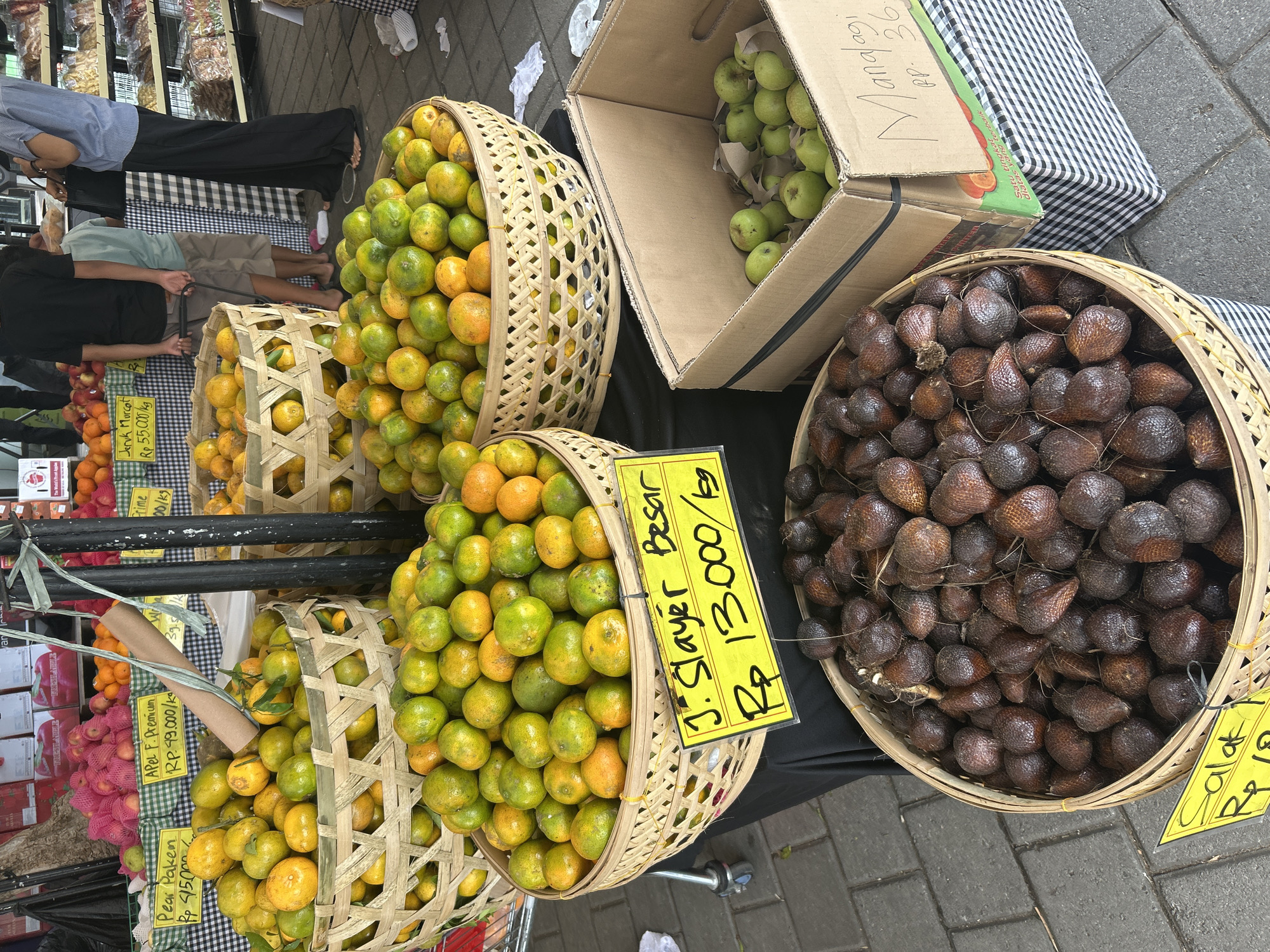 This image depicts a vibrant fruit stand at the Sukawati Art Market in Sukawati, Indonesia. The market appears busy with various fruits displayed in woven baskets and cardboard boxes. In the foreground, a large woven basket is filled with salak, also known as snake fruit, which are small, brown, and scaly. A yellow sign below this basket reads "Salak Rp 18,-/kg". To the right of the salak, a cardboard box contains green apples. A label on this box, written in Indonesian, indicates "Manalagi" and "Rp. 36,-".
Behind the salak, another large woven basket is filled with oranges and tangerines, a mix of green and orange hues. A prominent yellow sign attached to this basket reads "J. Slayer Besar Rp 13,000/kg". Further behind, more baskets overflow with oranges, and some red apples are visible in the background.
Several people are present at the market. In the background, a person wearing a black t-shirt and shorts is seen holding a leash connected to a dog, and another person in a blue collared shirt and black pants stands near a display of packaged goods. The ground is paved with interlocking stones, and the market stall is covered with a black fabric. The overall scene is one of a bustling outdoor market showcasing fresh produce.