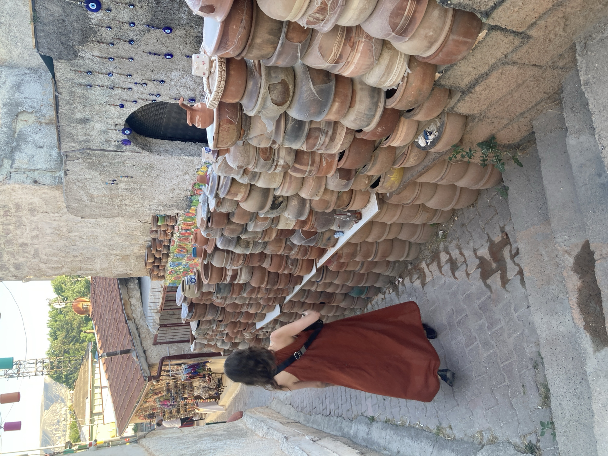 The image depicts a narrow, stone-paved street in Avanos, Turkey, lined with a shop displaying a large collection of terracotta pots and other decorative items. A woman in a long, burnt-orange dress walks away from the camera down the street. She carries a black crossbody bag.
The shop's facade is adorned with hundreds of stacked terracotta pots, many of which are partially covered in plastic sheeting. Some pots have small decorative items like cups attached. Above the pots, strings of blue evil eye charms are hung against a stone wall, with an arched doorway visible in the background.
Further down the street, more stalls are visible, displaying a variety of goods, including colorful hanging lamps and what appear to be textiles or scarves. Beyond the immediate shops, rooftops and trees are visible, with a hazy mountain landscape in the distance, suggesting a warm, possibly sunny day. The overall impression is of a bustling marketplace in a historic Mediterranean town.