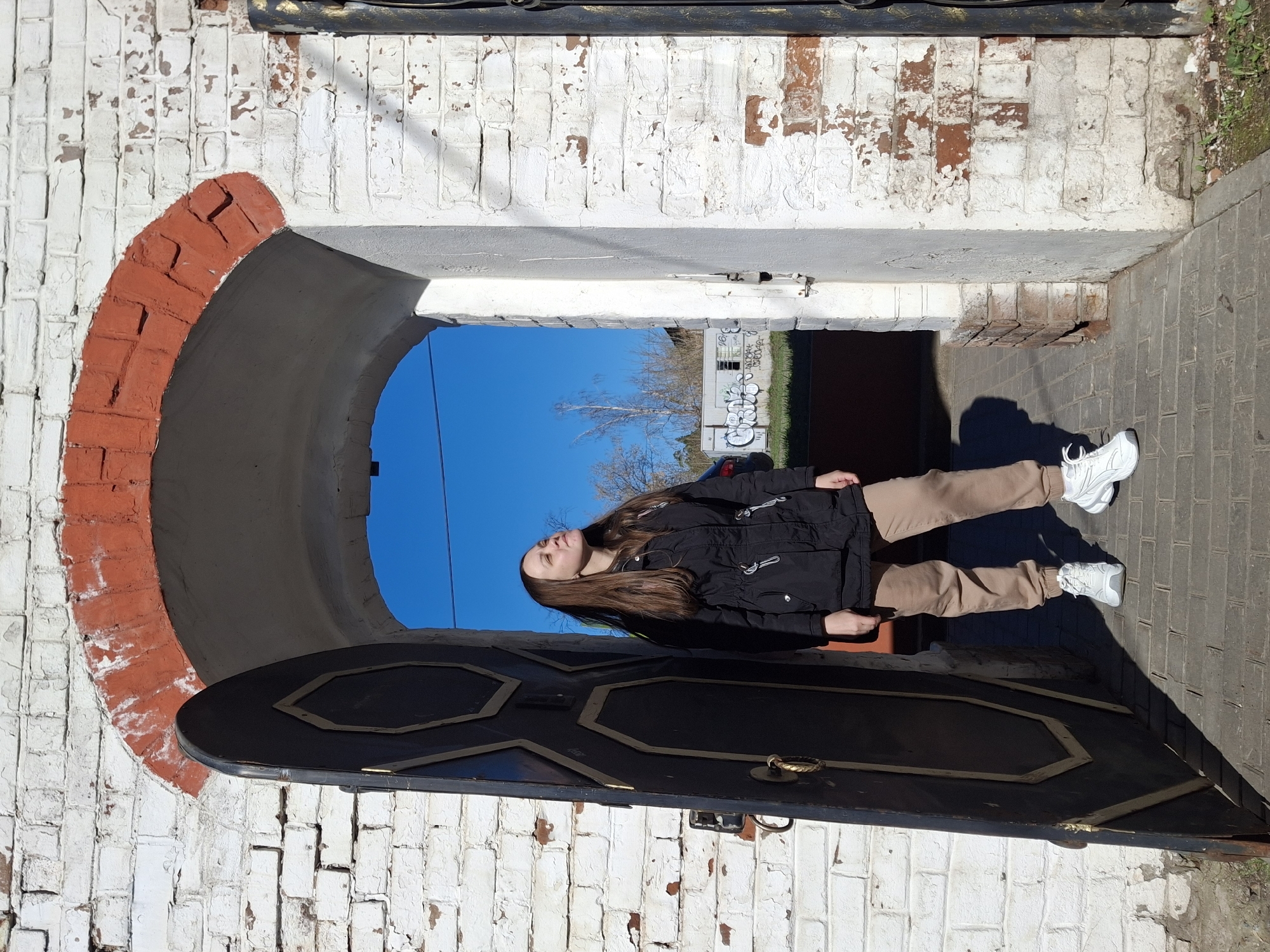 A girl stands in the open doorway of a brick building under a blue sky. The doorway features a red brick arch. The girl is wearing a black jacket and tan pants with white sneakers. Beyond the doorway is a building with graffiti. The location is Sarapul, Russia.
