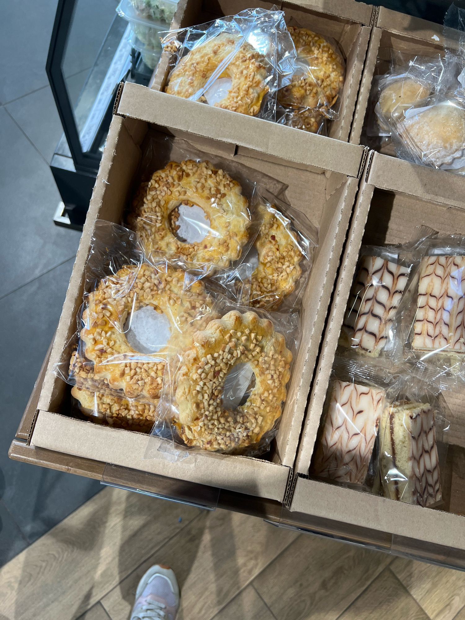 The image displays a close-up, overhead view of a retail display case filled with individually packaged pastries. The display is divided into cardboard compartments. In the foreground, a compartment contains several ring-shaped cookies coated in chopped nuts. Behind these, another compartment holds similar nut-coated cookies. To the right, two compartments contain rectangular pastries with a marbled frosting pattern. The background shows a glass display case with a blurry salad in a plastic container and more pastries, suggesting this is a bakery or food service establishment. The floor appears to be wooden laminate. A person's foot wearing a light pink and white sneaker is visible in the lower right corner of the frame. The scene is well-lit, highlighting the textures and colors of the baked goods.