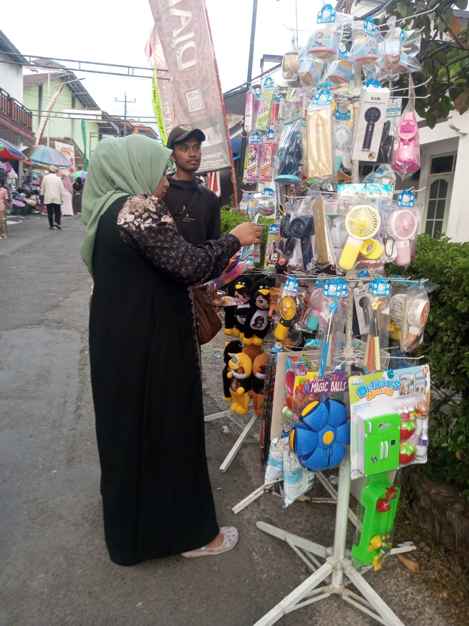 In Pandansari, Indonesia, a person wearing a hijab and a black abaya with patterned sleeves is browsing a street vendor's stall. The vendor's stall displays a variety of merchandise, including small fans, plush toys, and toy kitchen sets. A young man in a black t-shirt and a baseball cap stands behind the stall, observing. The scene appears to be on an asphalt street, with buildings and lush greenery visible in the background. The sky is overcast.