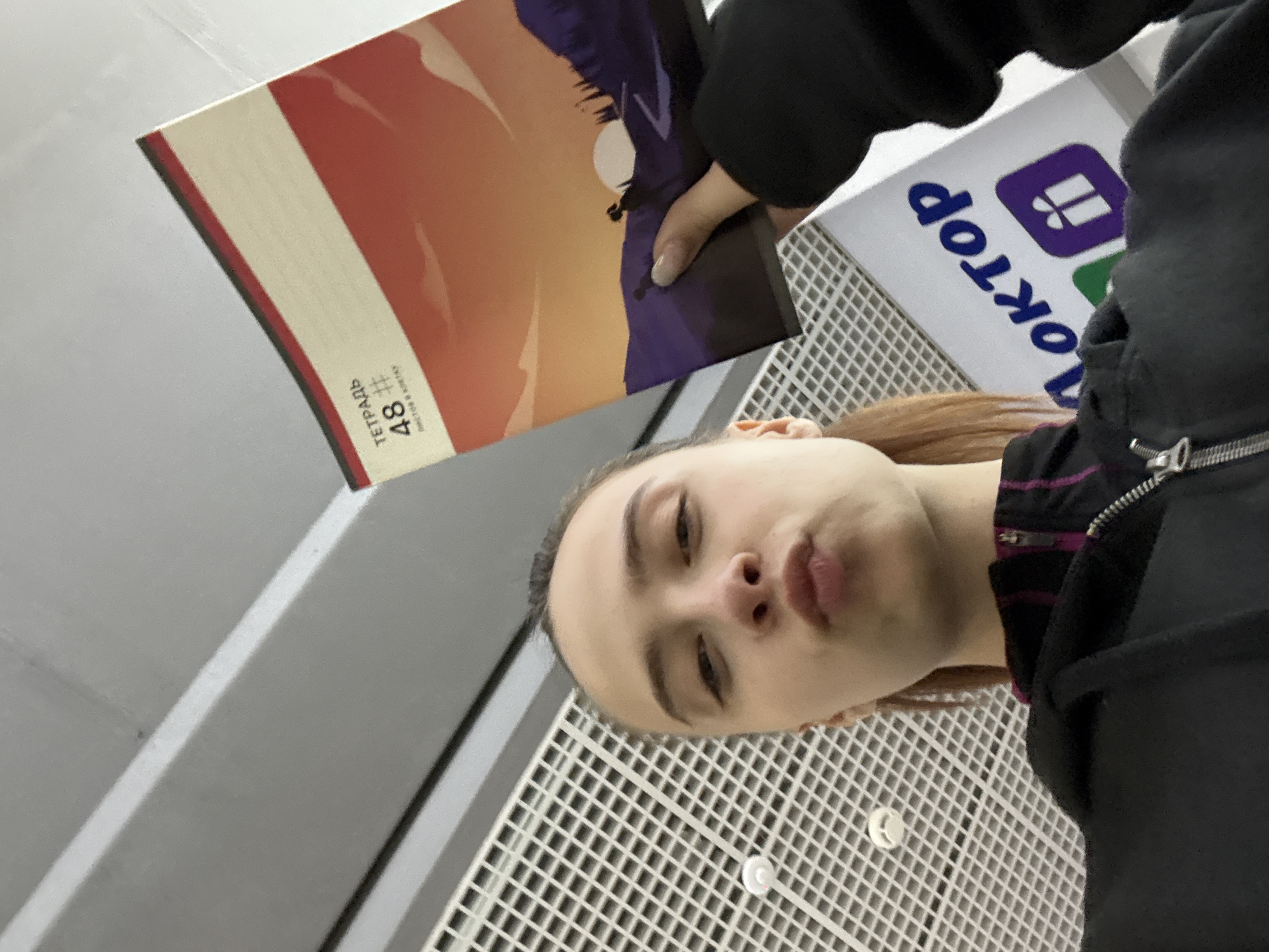 The image shows a person indoors, possibly at a bookstore called "Читай-город" in Chaikovsky, Russia, as a sign with the store's logo is visible. The person is holding a notebook with a cover depicting a landscape with orange and purple tones, and a notebook with 48 sheets. The person is wearing a black jacket and has light brown hair. The background includes a grid-patterned surface, and parts of a wall. The image appears to be a selfie or taken from a low angle.