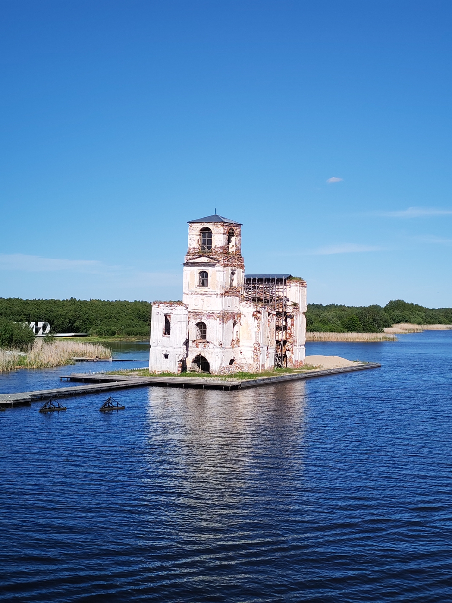 This image captures the poignant ruins of the Church of the Nativity of Christ, located in Krokhino, Russia, standing majestically within a serene body of water. The scene is dominated by the tall, decaying structure, primarily white with large sections of exposed red brickwork where plaster has eroded. The church features a prominent bell tower, topped with a dark, conical roof, and numerous empty arched windows. Extensive damage is visible on the main body of the building, with broken walls and internal wooden scaffolding or supports exposed on its right side.
The ruined church rests on a small, sandy patch of land amidst the dark blue, rippling water, which appears to be a lake or reservoir. Wooden jetties and walkways extend across the water, providing access to the church's base from the left shore. Near these walkways, two dark, triangular structures are partially submerged.
In the background, a dense, verdant forest lines the distant banks of the water body. The sky above is a brilliant, clear blue with only a single, tiny white cloud visible high up, indicating bright, sunny weather, likely midday or early afternoon. Tall reeds and grasses grow along the nearer shoreline to the left.
No people are visible, and no specific activity or event is taking place, contributing to a sense of historical solitude and quiet contemplation. There is no visible text in the image. The overall impression is one of enduring history against a backdrop of natural beauty and decay.