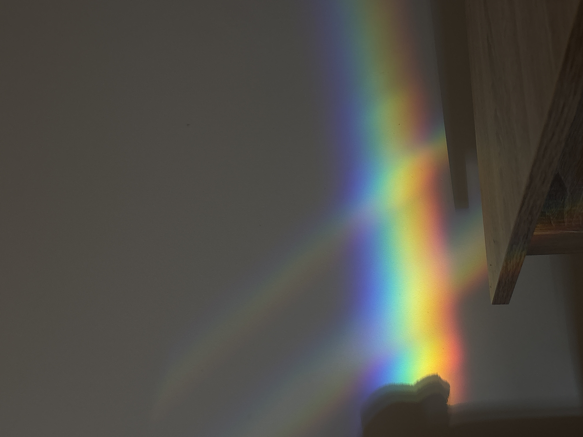 The image shows a rainbow spectrum cast on a light gray wall in what appears to be an interior space. A portion of a wooden structure, possibly shelving or a frame, is visible on the right side of the frame. The rainbow's colours range from violet and blue through green, yellow, orange, and red. A shadow, possibly of an object or person, is visible at the bottom of the rainbow spectrum. The location of Belgrade, Serbia, is not discernable from the information in the image.