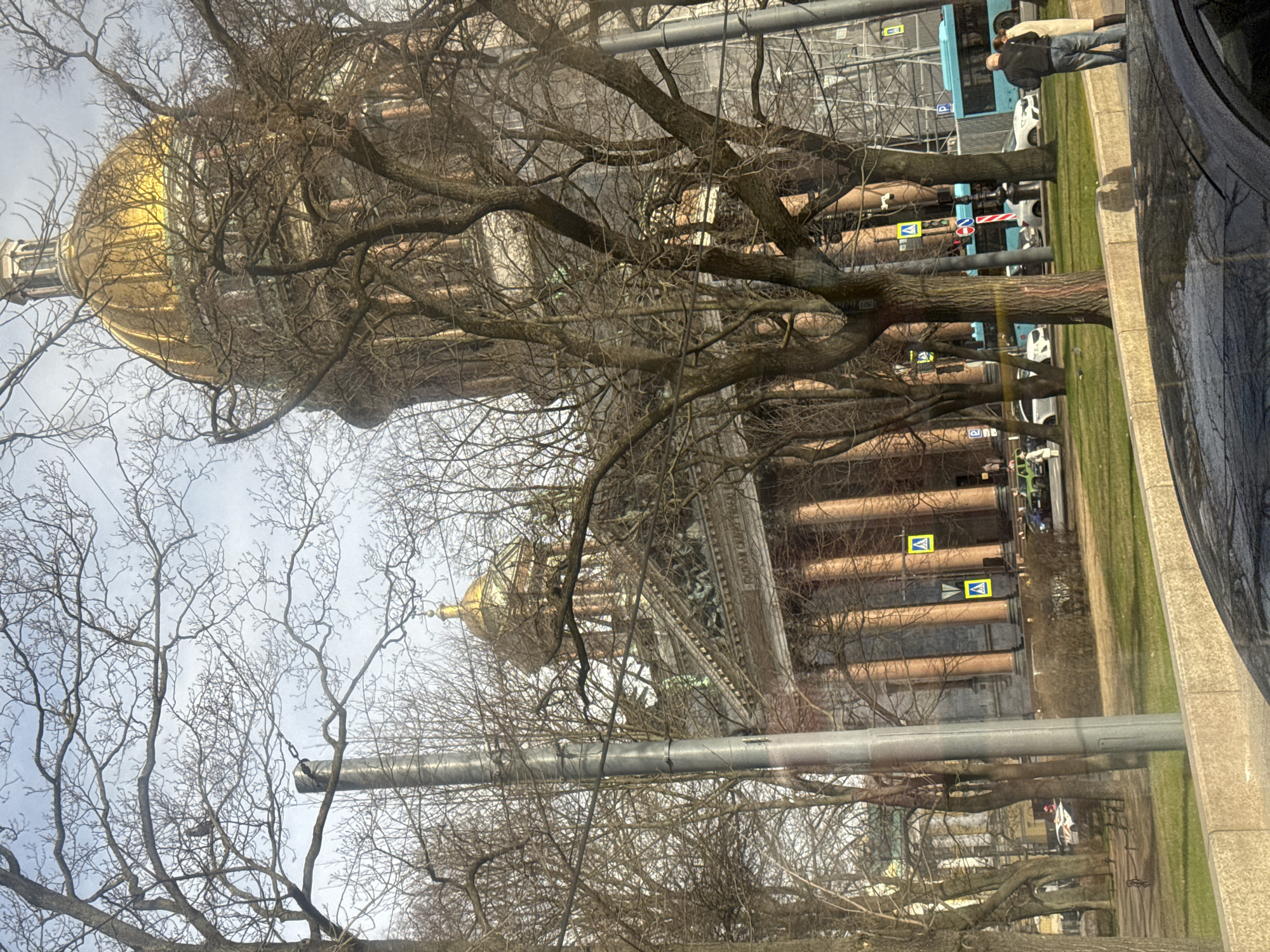 The image shows Saint Isaac's Cathedral in Saint Petersburg, Russia, seen through bare tree branches and a window. The cathedral's gold dome is prominent. The image includes a view of the street with parked cars and pedestrians. Also visible are pedestrian crosswalk signs mounted on the building's exterior. The sky is overcast.