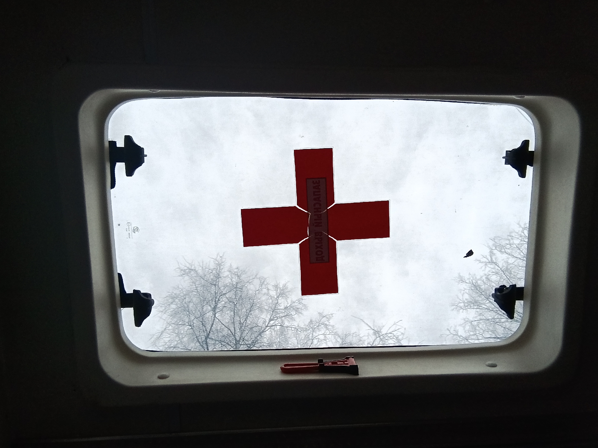 From inside a vehicle, a roof hatch window reveals a cloudy, gray sky and bare tree branches covered in frost. A red sticker in the shape of a plus sign ("+") is affixed to the window’s interior, with Cyrillic text vertically oriented on the central vertical bar. Four black latches are positioned at the corners of the window frame. A small red and black object lies on the frame at the bottom of the window. The exact location in the city of Chaikovsky, Russia is unknown.