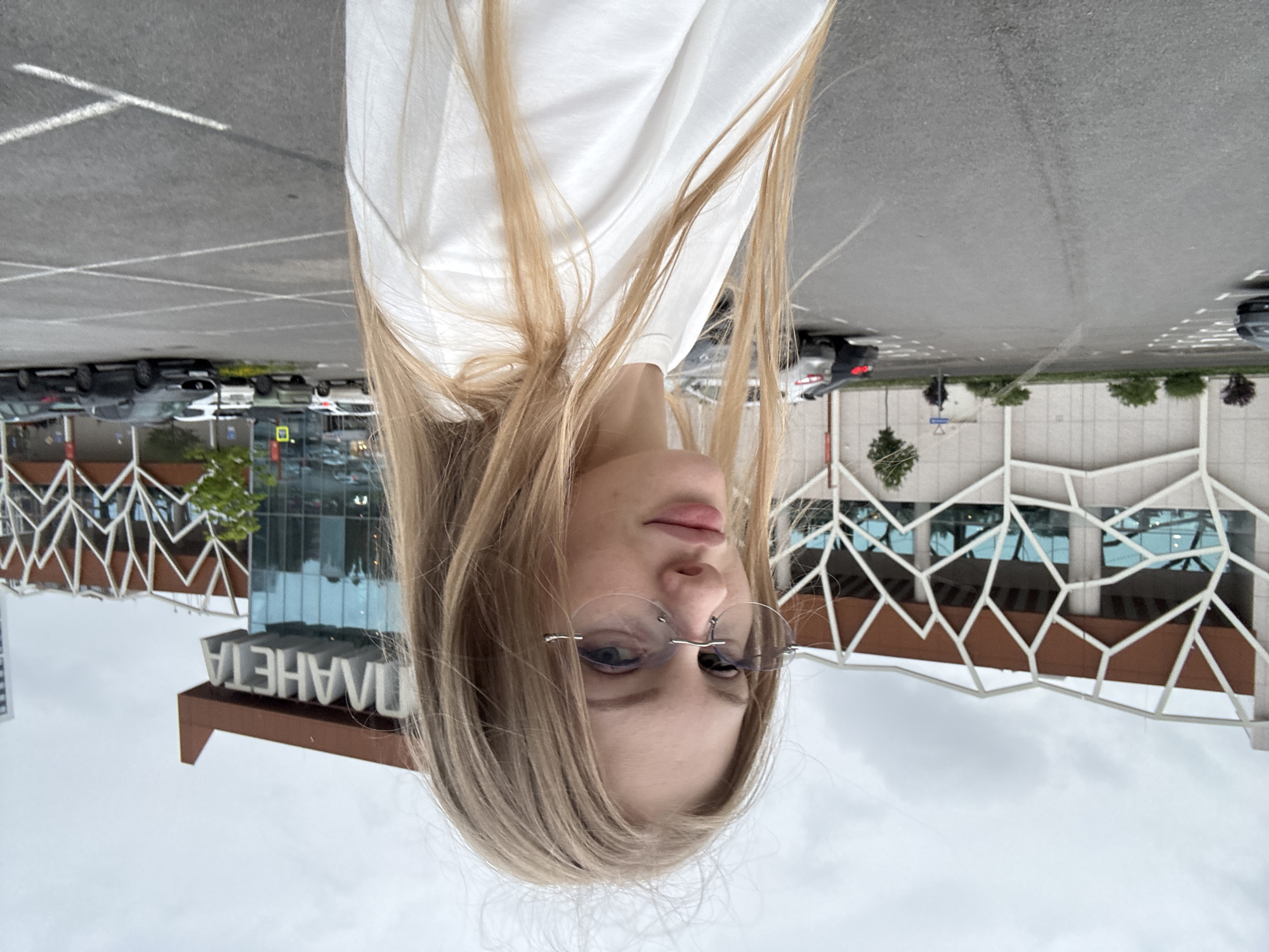 A young, blonde woman with light skin and light blue eyes, wearing rimless glasses and a white t-shirt, is photographed outdoors in a parking lot. She is looking towards the camera with a neutral expression. In the background, a modern shopping mall with a distinctive geometric facade and the word "ПЛАНЕТА" (Planeta) prominently displayed is visible. The sky is overcast. Several cars are parked in the lot. The location is identified as the "Planeta" shopping and entertainment center in Perm, Russia.