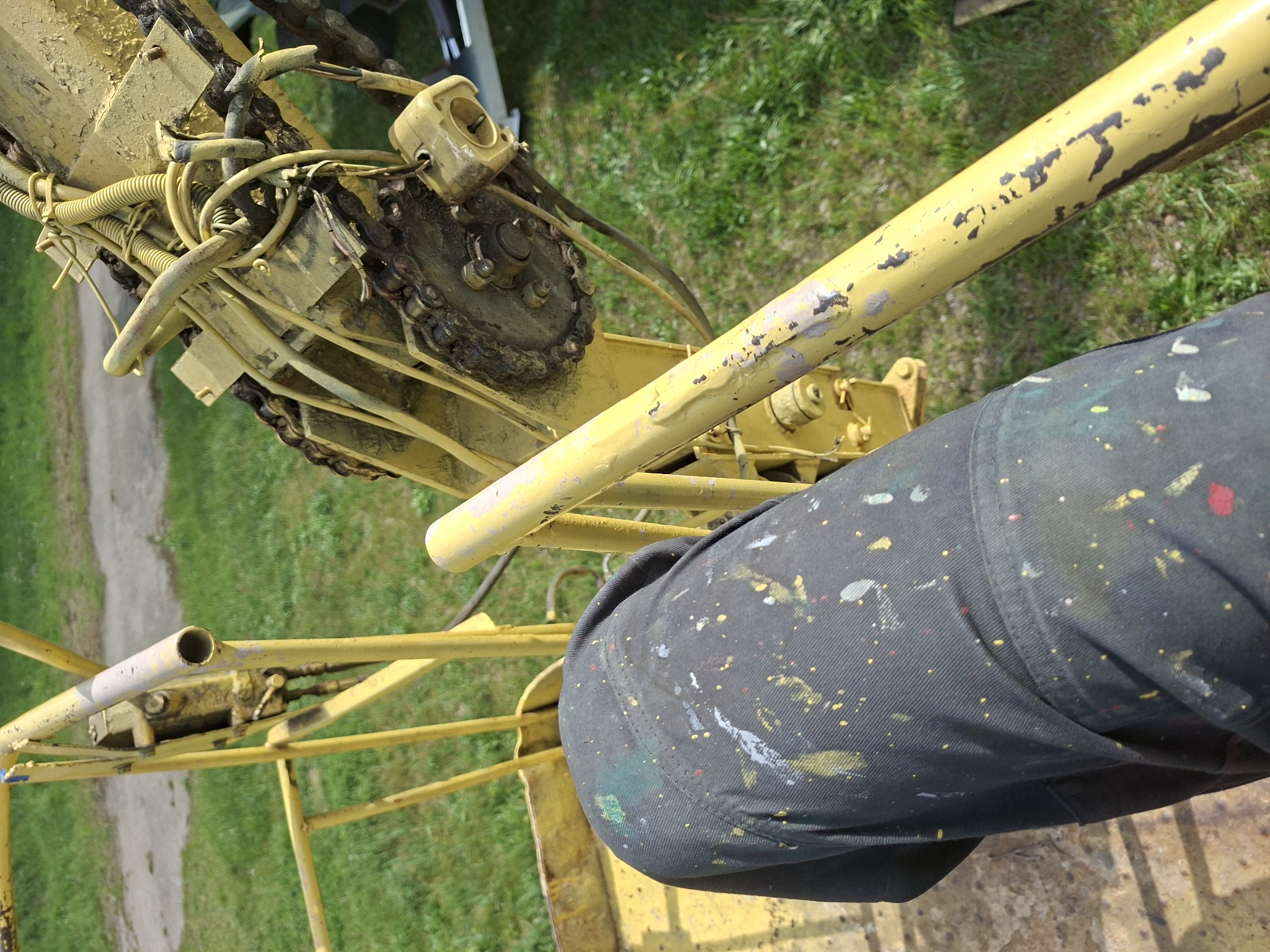 A ground-level, high-angle, outdoor shot shows a yellow, mechanical lift in an area with a grassy and dirt ground cover, ostensibly in the city of Zhabinka, Belarus. The lift’s structure is primarily tubular, with peeling yellow paint. The mechanical components, including a chain and various cables and hoses, are also painted yellow. A leg covered in black pants with paint splatters is positioned to the right, near the lift’s support. The background is a mix of green grass and a dirt pathway.