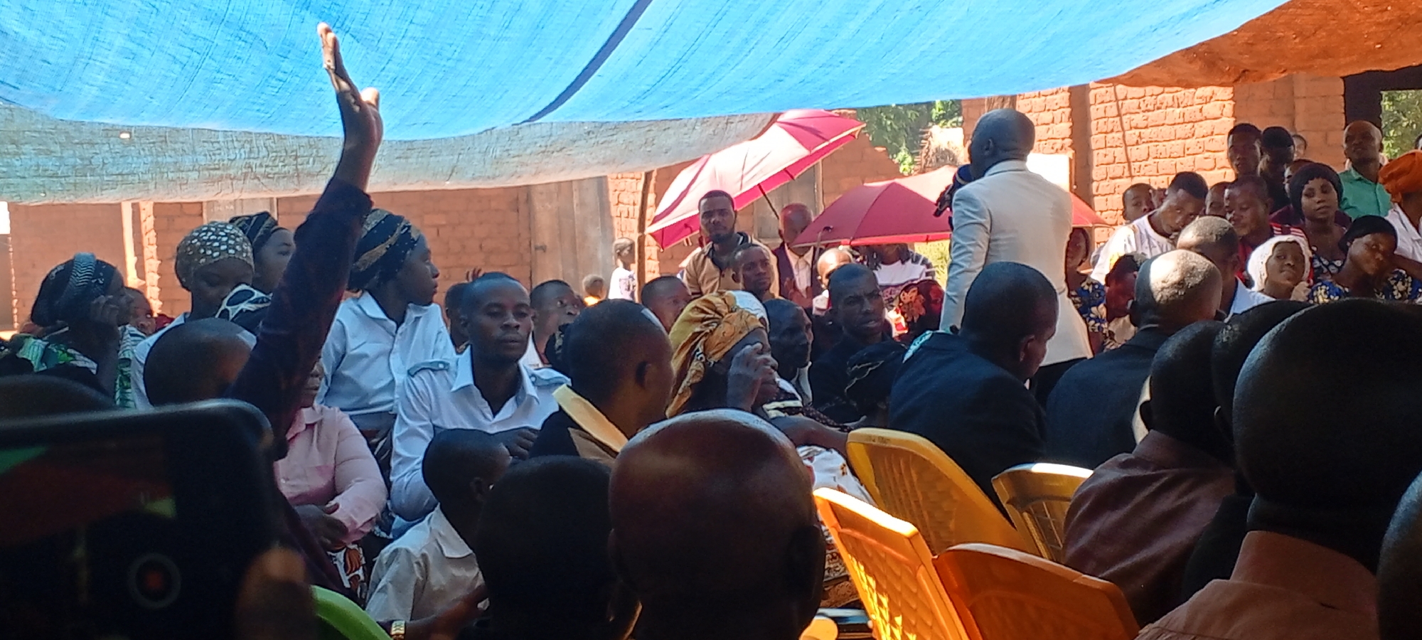A medium shot shows a gathering of people under a large blue tarp in what appears to be an outdoor event in Kalemie, Democratic Republic of the Congo. The location seems to be a yard with a brick building in the background.
The majority of the people are seated on yellow plastic chairs, facing a man in a light-colored suit who appears to be speaking into a microphone. A person with an arm raised is standing on the left, possibly signaling a question or request. Several people are wearing traditional head coverings. An open red umbrella is visible further back in the crowd. A mobile phone can be seen in the lower left corner, indicating someone is capturing the event. The environment appears bright and sunny.
