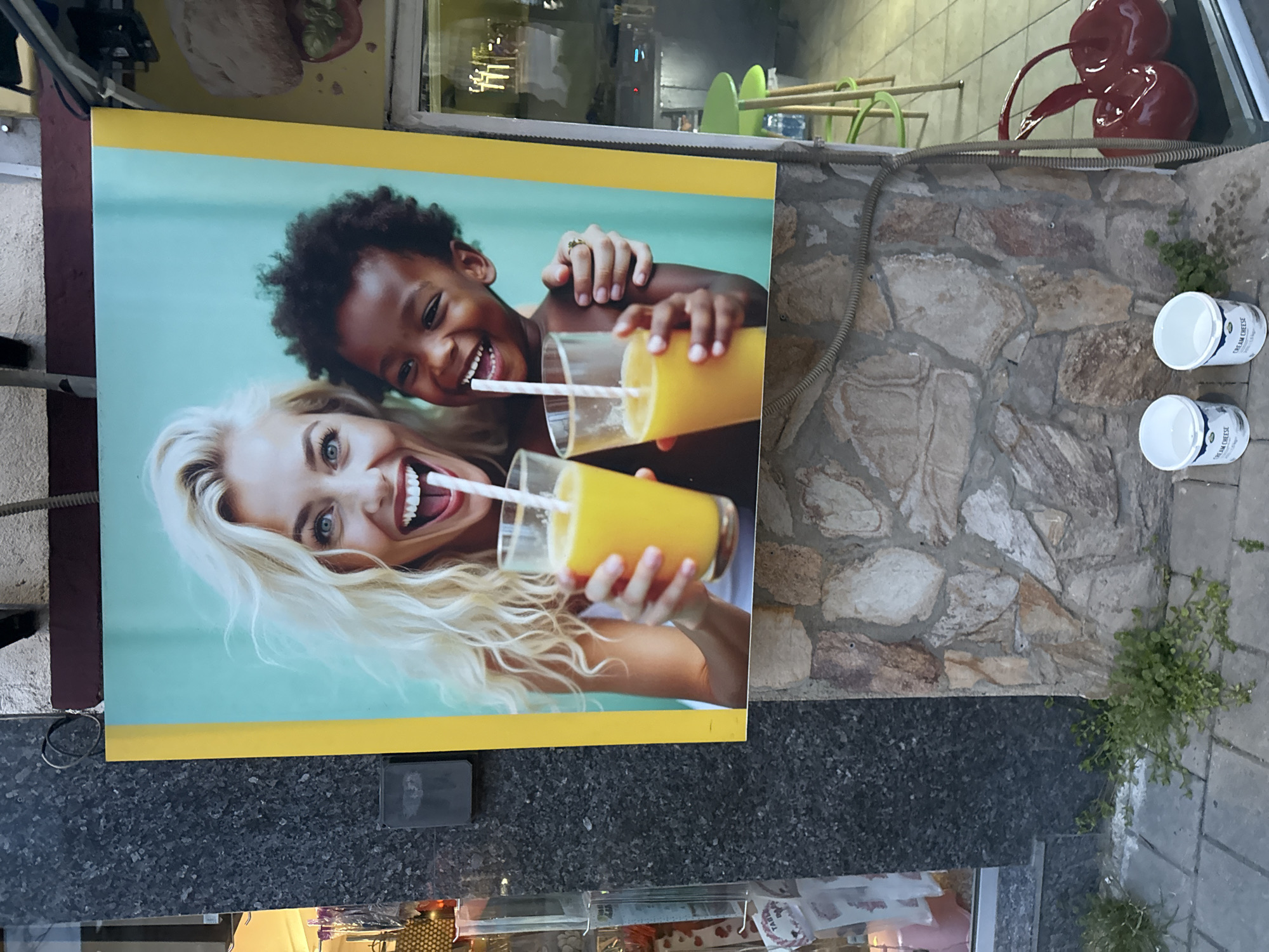 The image captures an outdoor advertisement in Varna, Bulgaria. The poster features a smiling woman with blonde hair and a young child of African descent, both drinking orange juice from glasses with straws. The background is a light turquoise color, and the poster has a yellow border. The advertisement is affixed to a textured wall constructed from various shades of tan stone. Adjacent to the wall, there is an area with foliage and two white tubs with the label "Cream Cheese". Above, a red cherry decoration is visible. In the background, the blurred outlines of tables and chairs suggest a cafe or restaurant setting.