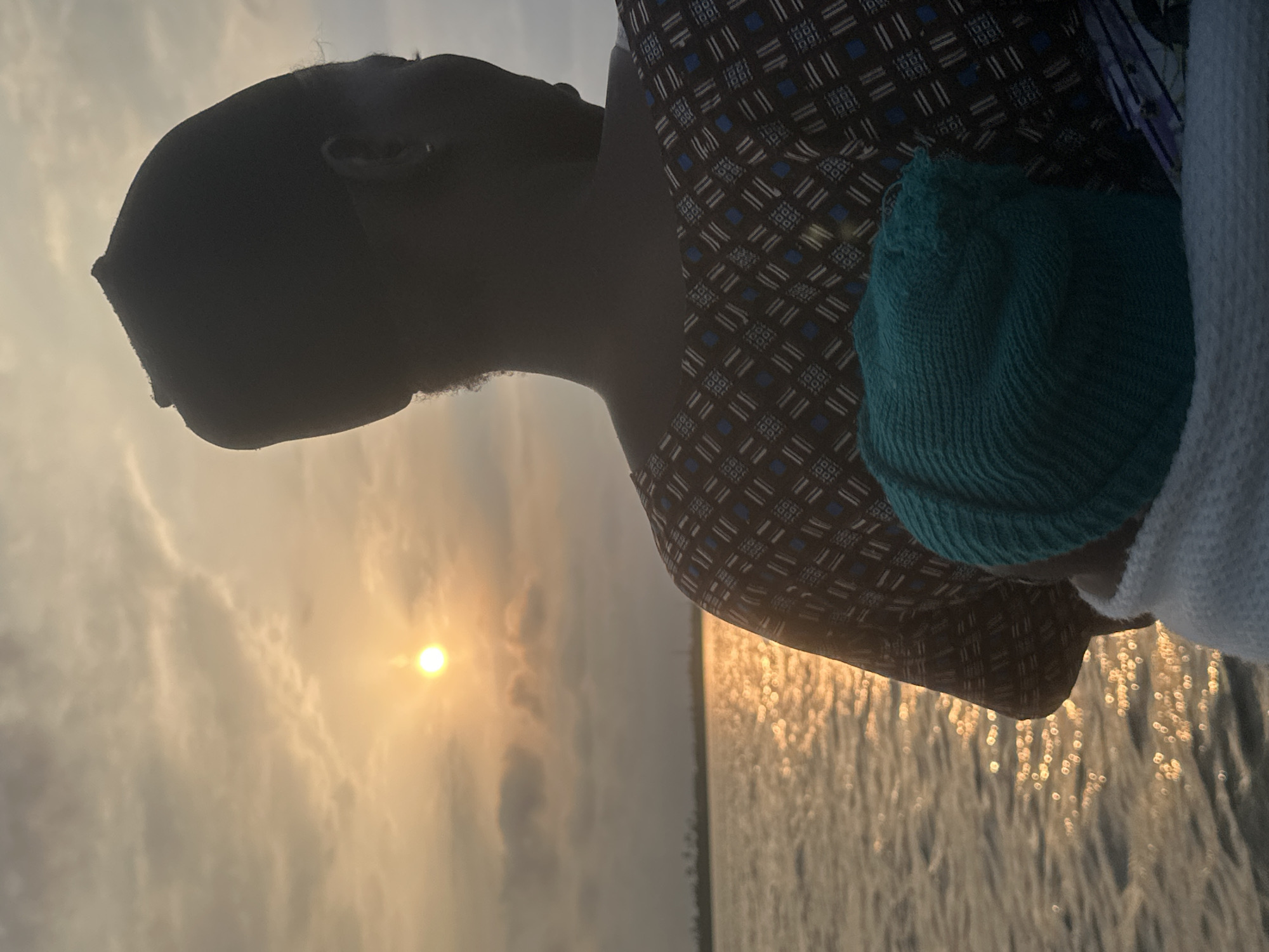The image depicts a person, possibly a mother, seen from behind, carrying a baby wrapped in a teal knitted cap and a white blanket. They are silhouetted against a sunset over a body of water, likely a lagoon or the ocean, with a distant shoreline visible. The sun, a bright orb with rays, is setting amidst scattered clouds, casting a warm, golden glow on the water. The person is wearing a dark patterned shirt and a dark cap. The setting appears to be along the coast of Badagry, Nigeria, known for its historical significance and proximity to the Lagos Lagoon and the Atlantic Ocean, where sunsets over water are common.
