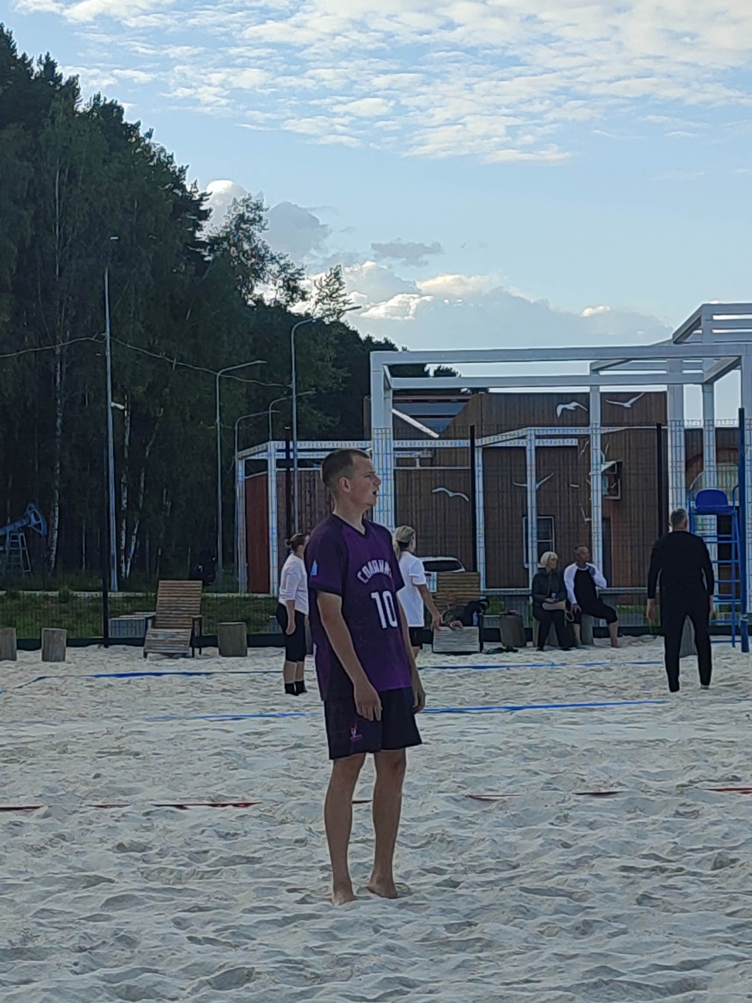 The image shows a beach volleyball court on a sandy area, with a line of trees in the background. The sky is partly cloudy, suggesting it is daytime, possibly late afternoon.
In the foreground, a young man wearing a purple t-shirt with the number "10" and dark shorts is standing on the sand, looking to his right. His expression appears neutral. Behind him, several other people are visible. To the left, a woman in a white shirt and dark shorts is standing. Further back, some people are sitting and standing near a wooden structure with a roof. To the right, a person in a black outfit is standing and facing away from the camera. A blue ladder and a chair are part of the volleyball court setup.
The scene depicts a casual gathering or a sporting event, likely beach volleyball, taking place outdoors. The setting appears to be a recreational area, possibly a park or a beachside facility, given the presence of sand, volleyball equipment, and seating. The lighting and the presence of clouds suggest a mild, possibly breezy day. There is text visible on the t-shirt of the man in the foreground, which reads "COACHING" above the number "10".