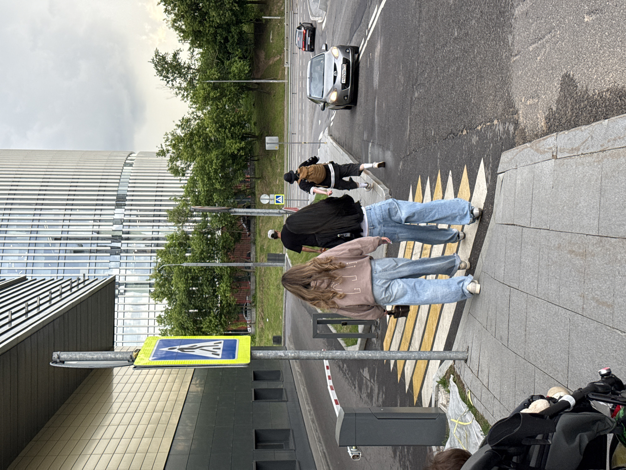 A pedestrian crossing is marked with yellow and white stripes on an asphalt road. Two women in loose-fitting light blue jeans and hoodies walk across the crossing. One woman has long brown wavy hair, and the other has long black hair. A man wearing a black t-shirt and a baseball cap walks behind them. Further down the road, a person with a backpack walks in the same direction. A silver car and a dark-colored car are on the road, with the silver car approaching the crossing. A modern building with a glass facade and ribbed vertical elements stands in the background, along with green trees. A pedestrian crossing sign with a blue background and a white pedestrian symbol is posted on a metal pole. A gate barrier is partially visible on the left side of the image. The sky is overcast.