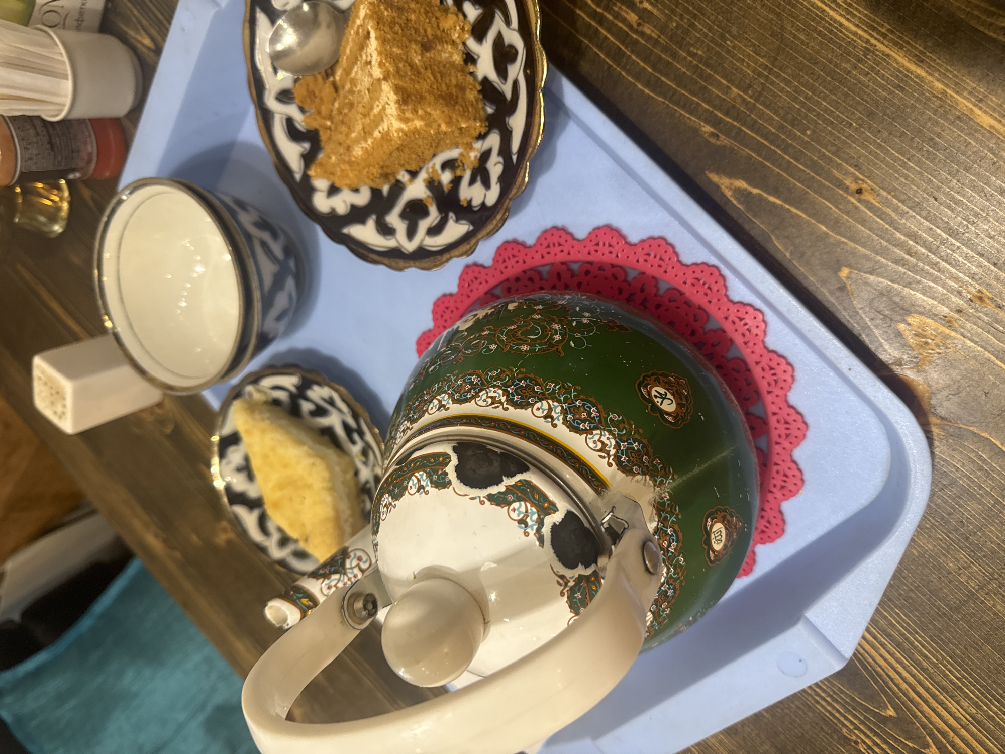The image depicts a tea set with two pieces of dessert on a light blue tray placed on a wooden table. The tea set includes a white and green patterned teapot with a white handle and lid knob, and a small white bowl with blue decorative trim. A pink doily is placed beneath the teapot. To the right of the teapot, on a dark blue and white patterned plate, sits a slice of layered cake, possibly a honey cake, with crumbs scattered around it. Another dark blue and white patterned plate to the left of the teapot holds a triangular piece of pastry, resembling baklava. In the background, out of focus, are what appear to be condiment holders and possibly a bottle of sauce. The setting is likely a cafe or restaurant in Komsomolsk-on-Amur, Russia, suggested by the potential for local pastries and tea service. The lighting is warm, casting shadows and highlighting the textures of the food and table.