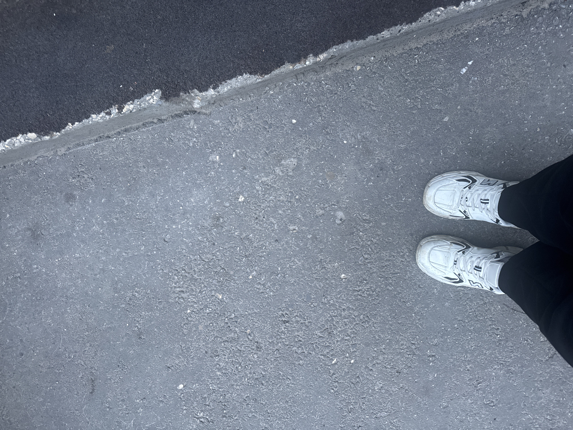 The image shows a person's feet wearing white sneakers and black pants, standing on a grey asphalt surface. To the right, there is a distinct edge where the grey asphalt meets a darker asphalt surface. The scene is not specific to Saratov, Russia, nor does it contain any details that would indicate this location. The lighting suggests it is daytime.