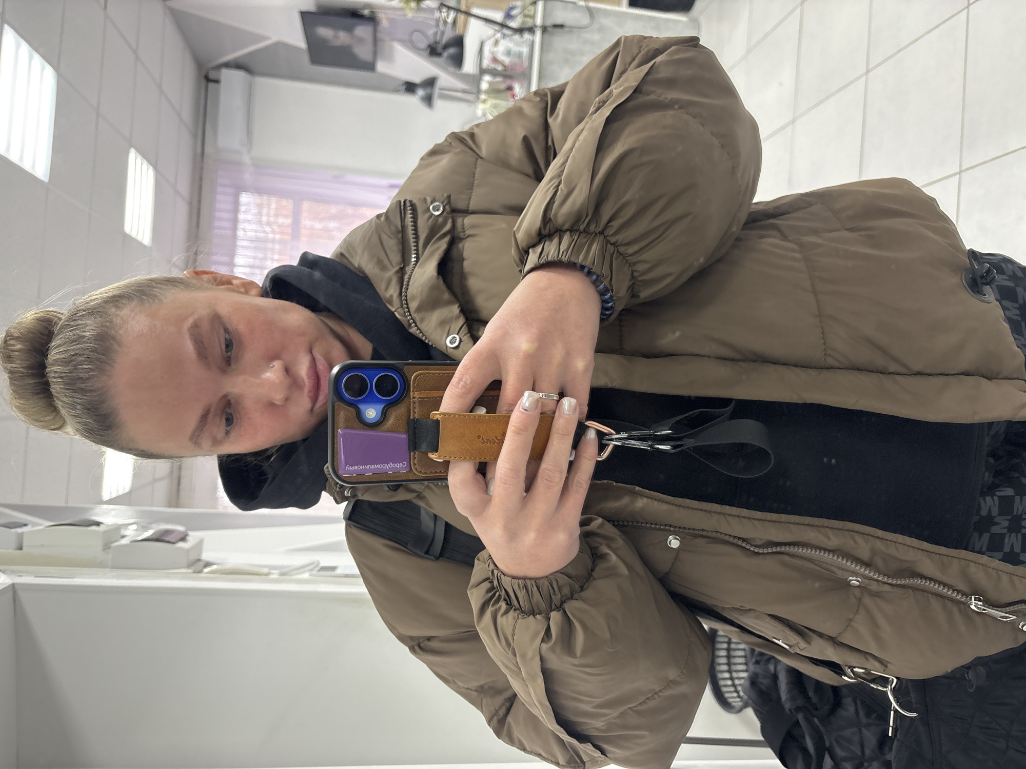 A woman with blonde hair in a bun is taking a selfie in a mirror. She is wearing a brown puffer jacket over a black hooded sweatshirt. She is holding a smartphone with a brown and purple phone case. The reflection in the mirror shows a tiled wall, a sink area with various items, and salon equipment. The woman appears to be in a salon in Perm, Russia, as indicated by the reversed inscription "Сделано во Владивостоке" (Made in Vladivostok) and the location of the inscription in the region of phone case color violet.