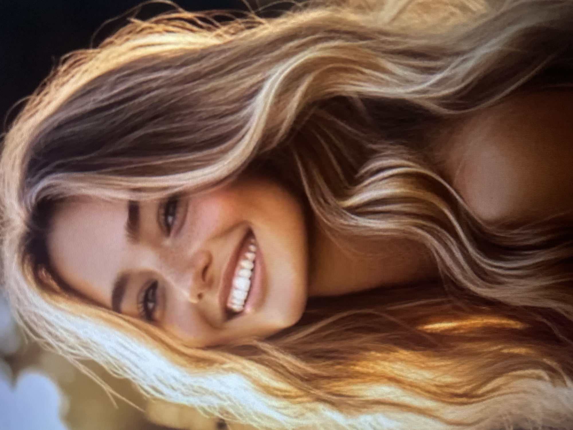 A close-up, medium shot of a young woman's face. She has long, wavy, blonde hair with highlights. She is smiling broadly, showing her teeth, and her eyes are looking towards the viewer. There are freckles visible on her nose and cheeks. The lighting is warm and suggests it might be sunset, with sunlight illuminating her hair and face from the side. The background is blurred, with hints of light and shadow. The image appears to be a portrait photograph. There is no indication of the city of Kołobrzeg, Poland within the image itself.