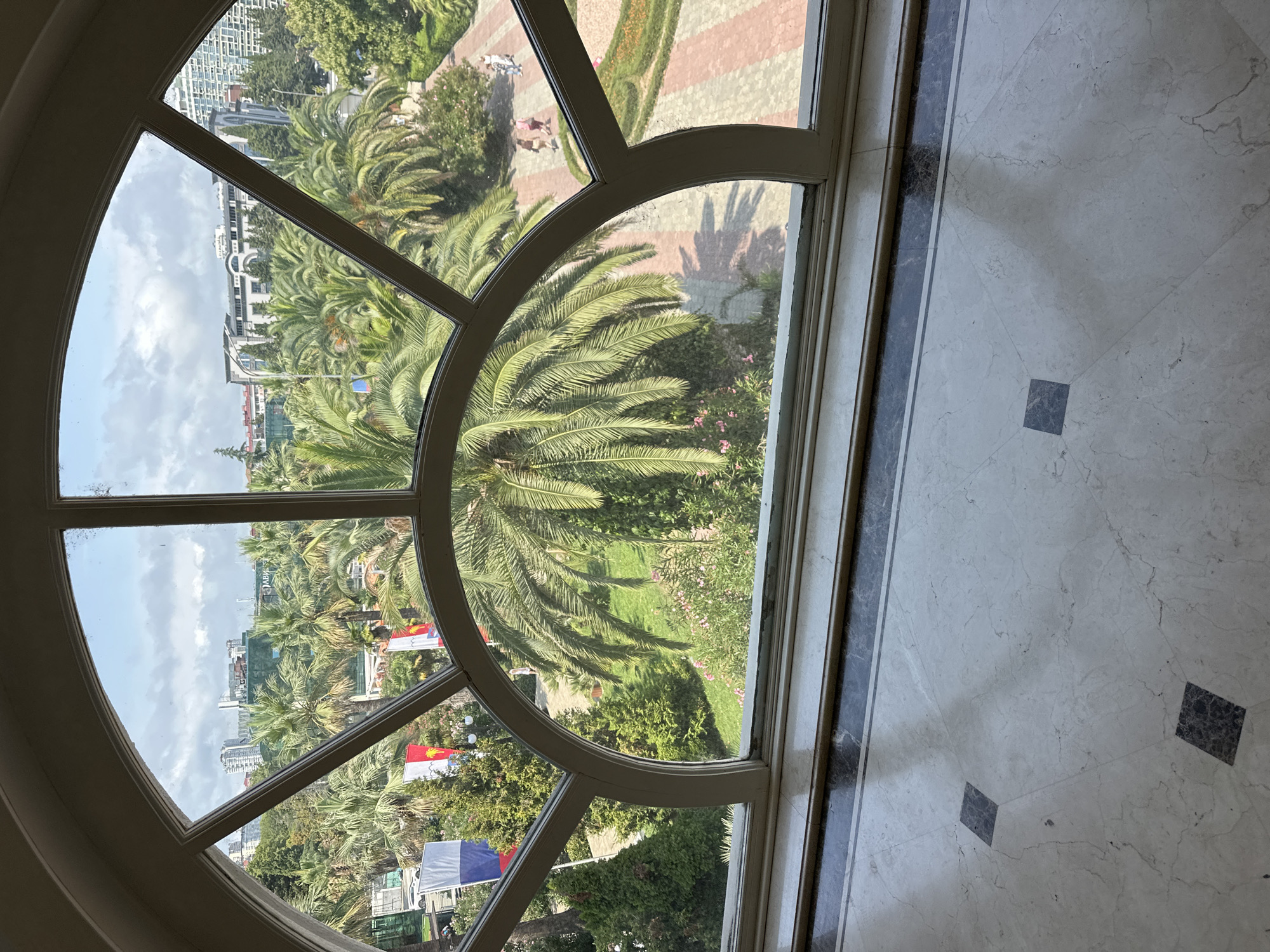 The image captures a view through a large, arched window with white wooden mullions, looking out onto a park-like setting in Sochi, Russia. The window frames a scene dominated by lush palm trees and other greenery. In the foreground of the outdoor view, a large palm tree with a full canopy is prominent. Beyond it, a paved walkway with red and light-colored stones runs through a manicured lawn. Several individuals are walking on this path, appearing as small figures in the distance. Further back, more palm trees and trees with darker green foliage are visible, interspersed with buildings. One building in the mid-ground appears to be a hotel or apartment complex with a light-colored facade and multiple windows. Flags, including what appears to be the Russian flag, are displayed on poles to the left of the pathway. The ground inside the room is a marble floor with a patterned inlay of dark squares. The overall impression is of a sunny day with scattered clouds in a city with a subtropical climate.