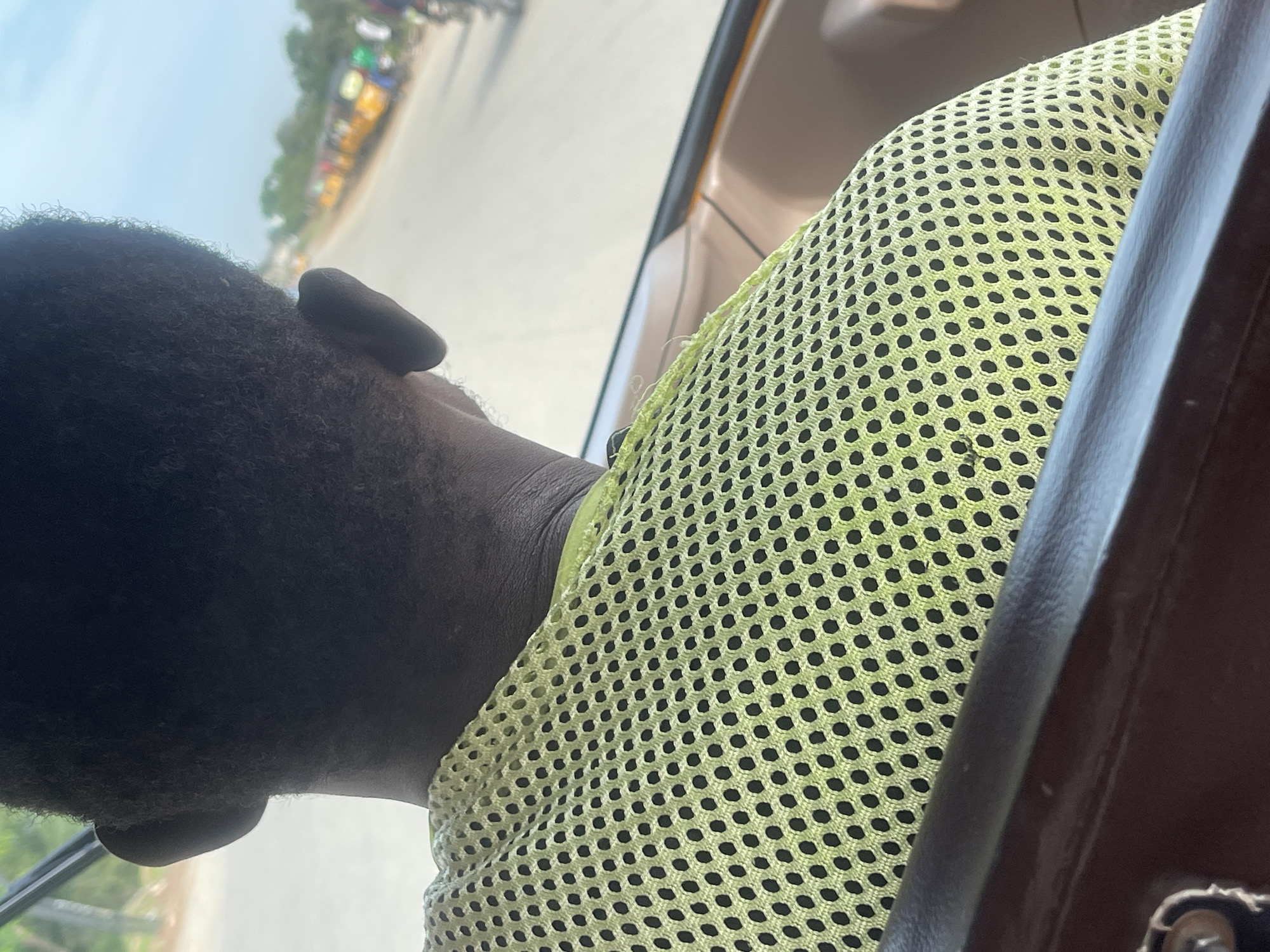 In Abuja, Nigeria, an overhead rear-view perspective captures an individual seated within a vehicle, looking forward onto a street. The subject, seen from behind, has dark, short, curly hair, with the right ear and nape of the neck clearly visible. The person is wearing a bright yellowish-green mesh shirt characterized by uniform black circular perforations. A dark brown, possibly leather or plastic, railing or frame of the vehicle runs horizontally across the lower portion of the image. Behind the subject's right shoulder, a lighter brown interior panel is discernible. Outside the vehicle, a light-colored road or street extends under a clear, light blue sky. A queue of distinctly yellow three-wheeled vehicles, consistent with "Keke Napep" tricycles prevalent in Nigeria, is visible along the right side of the road, either parked or in slow motion. Beyond these vehicles, green foliage outlines the distant horizon, interspersed with blurry indications of further human or vehicular presence, suggesting an active urban environment.