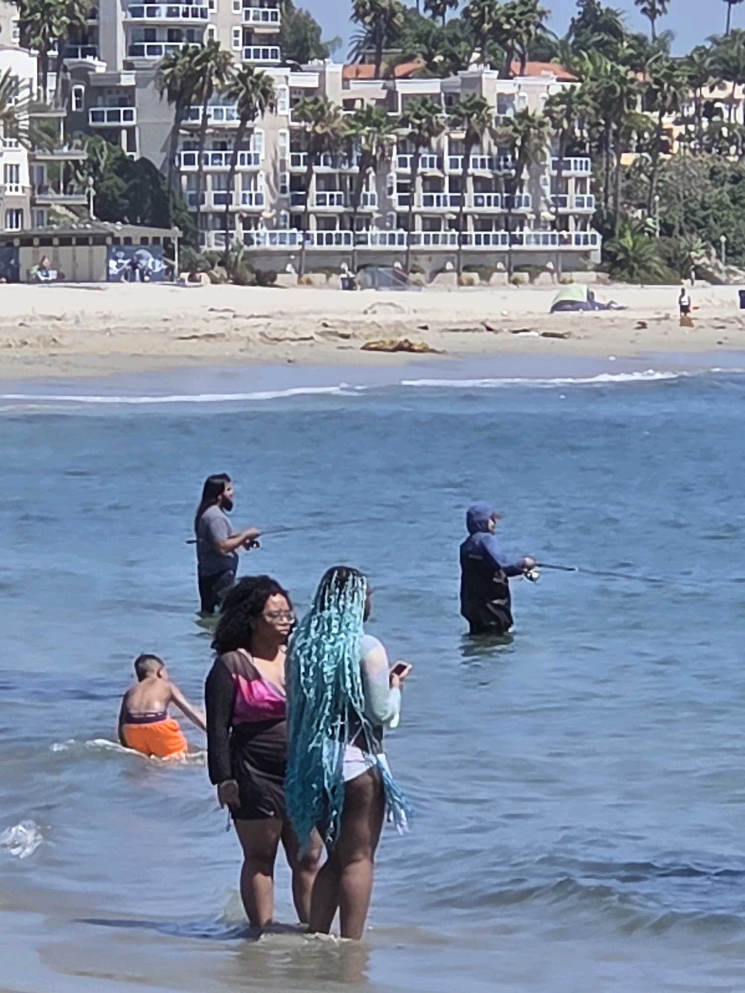 In Long Beach, California, a group of people are enjoying a sunny day at the beach. In the foreground, three individuals are wading in the shallow ocean water. The person on the far right has bright blue braids and is wearing a light-colored top and a white swimsuit. Next to them, a person with dark hair and glasses is wearing a black dress with pink accents. To the left of them, a young boy in orange swim trunks is standing in the water. In the middle ground, two men are fishing in the ocean. One man with long dark hair and a beard is wearing a gray t-shirt and dark pants. The other man is wearing a blue hooded jacket and dark waders. The background shows a sandy beach with palm trees and multi-story buildings. The ocean is calm with gentle waves breaking on the shore.