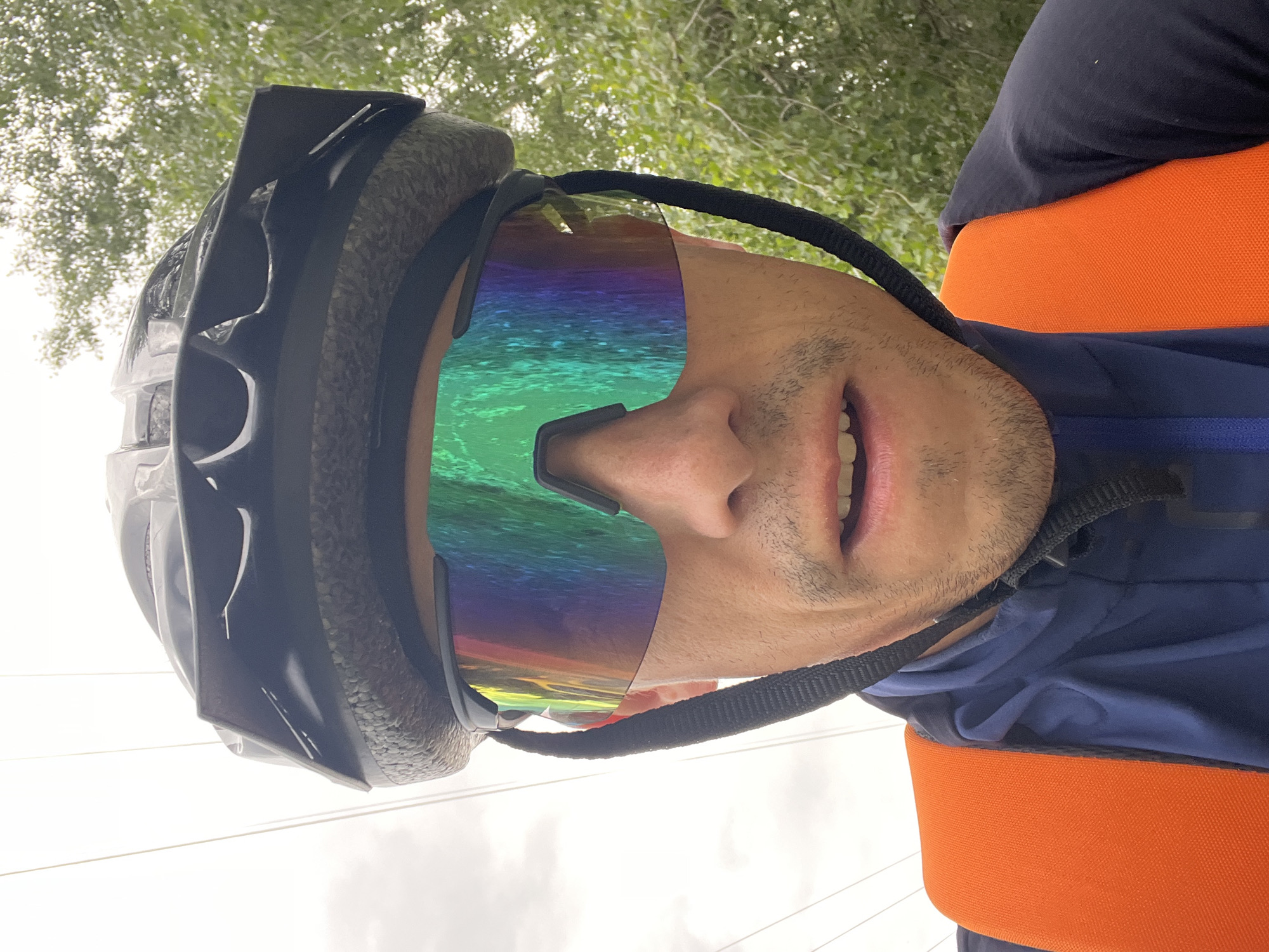 A close-up, low-angle, selfie-style photograph shows a person wearing a black cycling helmet and iridescent, wrap-around sunglasses that reflect a rainbow of colors. The person's face is partially obscured by the glasses, but their mouth is slightly open, revealing teeth. They are wearing a dark blue jacket and an orange vest over it. The background consists of green foliage and a cloudy sky, suggesting an outdoor setting, possibly near trees. The image does not contain any specific elements that would indicate the location as Chaikovsky, Russia.
