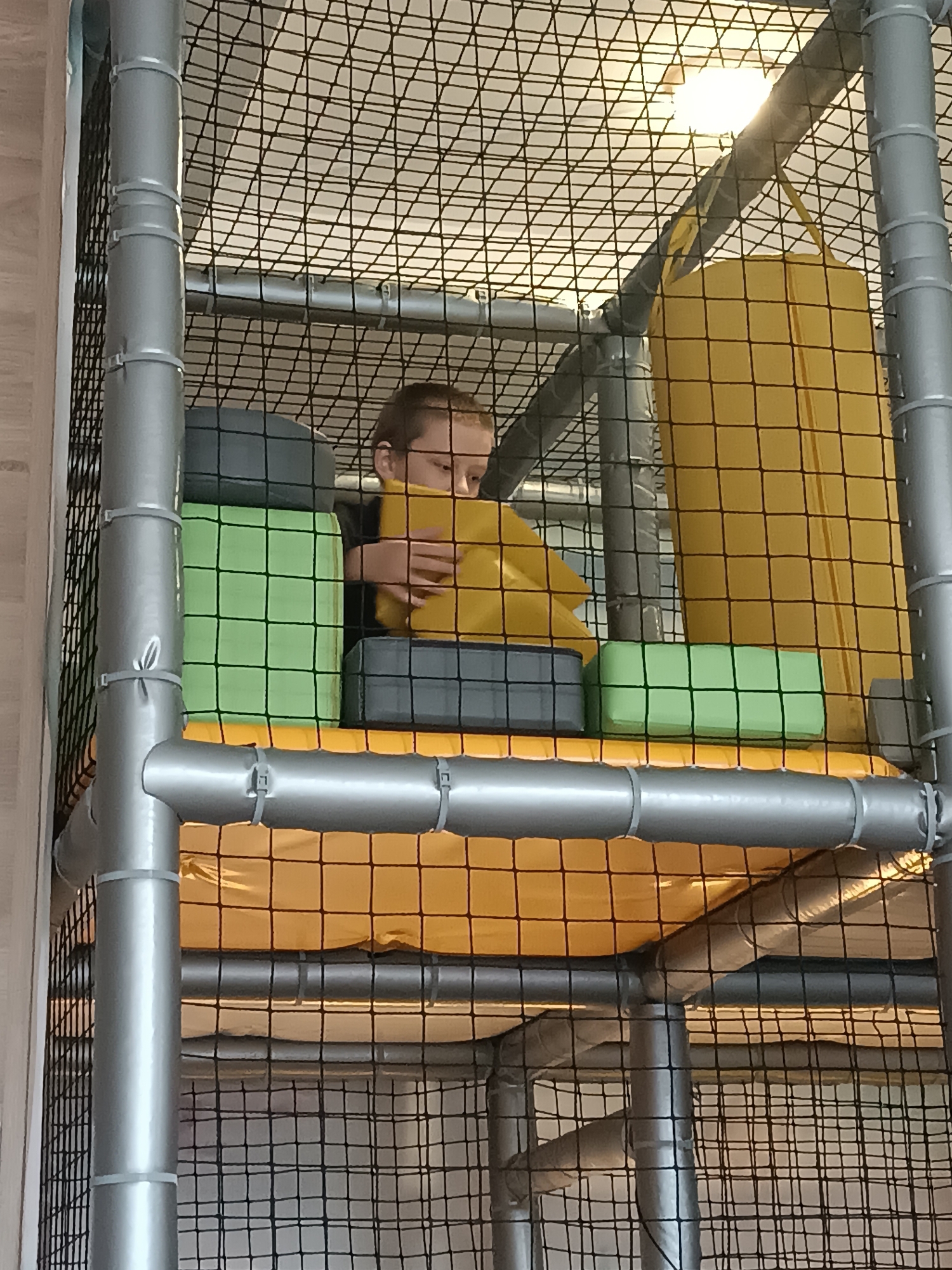 A child sits within a multi-level play structure. The structure is made of silver tubular poles and a black net enclosure. The child is surrounded by soft, rectangular blocks in colors of yellow, green, and grey. The child appears to be holding a yellow block. The background features a yellow padded floor. The location is unknown.