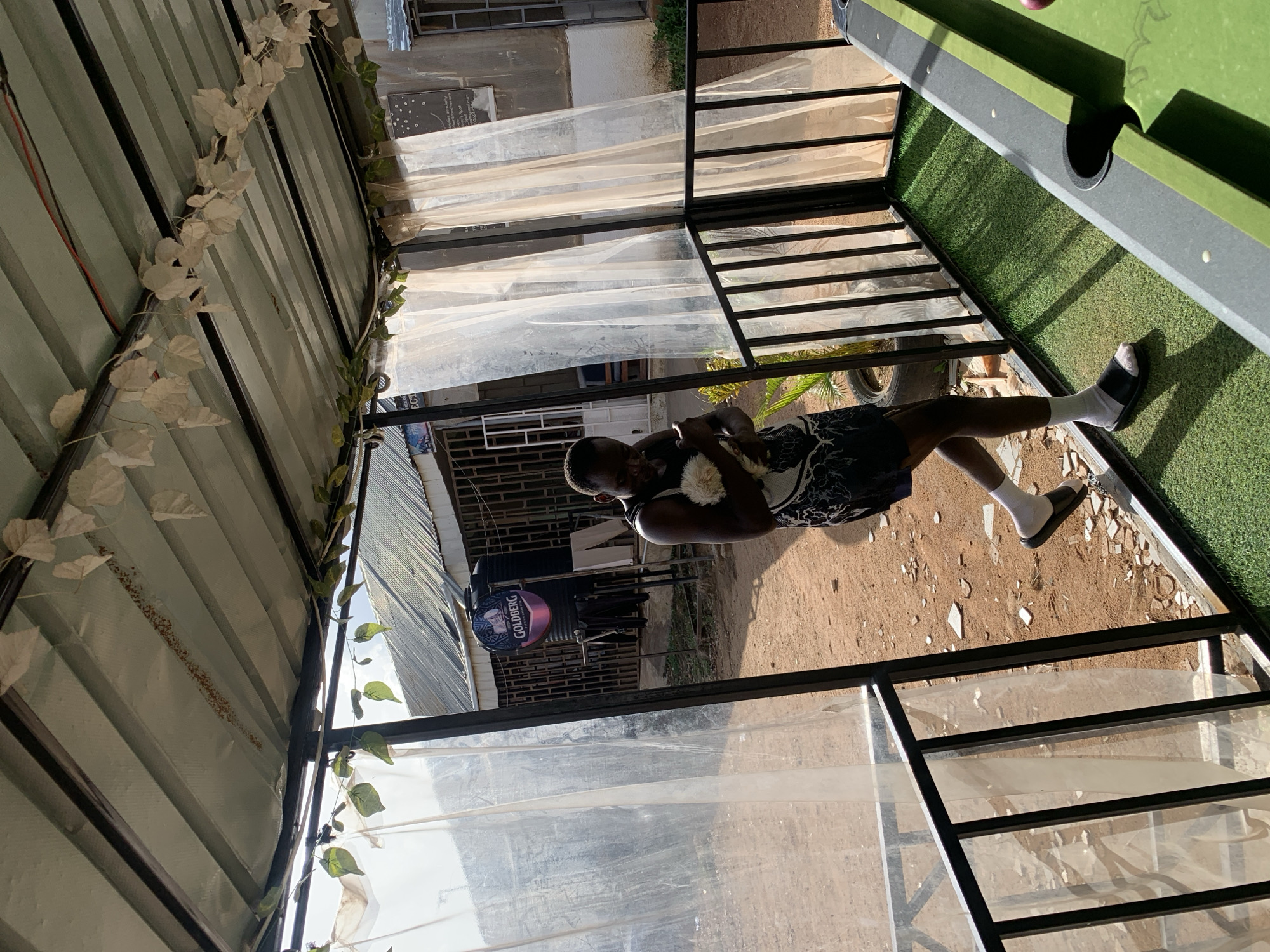The image depicts a man standing outside under a covered structure. He is wearing a patterned top and shorts, white socks, and sandals. The structure is decorated with white foliage and sheer white curtains. A pool table is visible to the right. In the background, buildings and a "GOLDBERG" sign can be seen. The location is Jos, Nigeria.