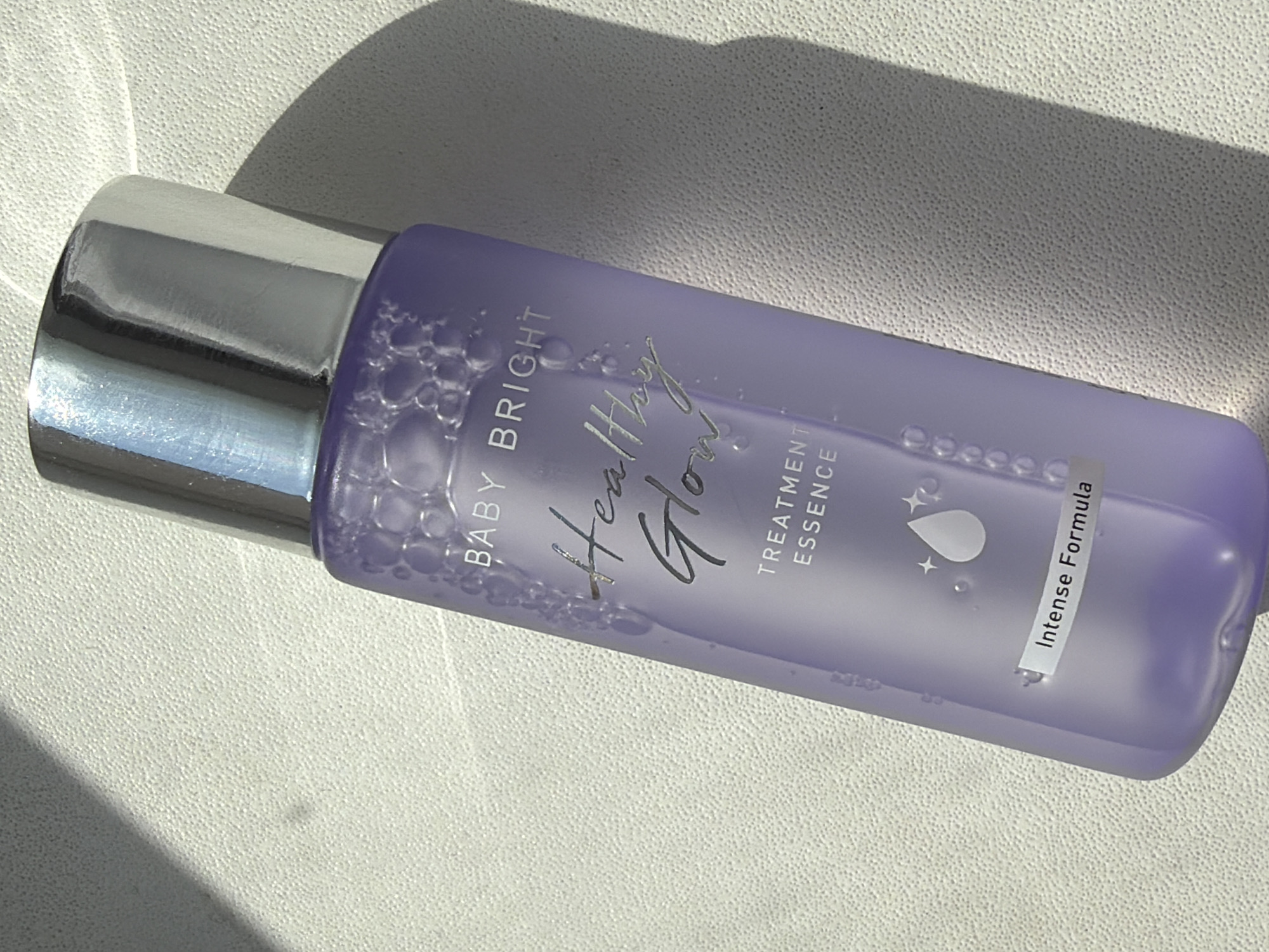 A horizontal eye-level shot depicts a partially filled translucent lavender bottle lying on its side. The bottle has a silver cap and is labeled "BABY BRIGHT Healthy Glow TREATMENT ESSENCE Intense Formula." The bottle is partially covered in what appear to be bubbles. It is resting on a textured white surface, which is illuminated by sunlight, creating a shadow above the bottle.
