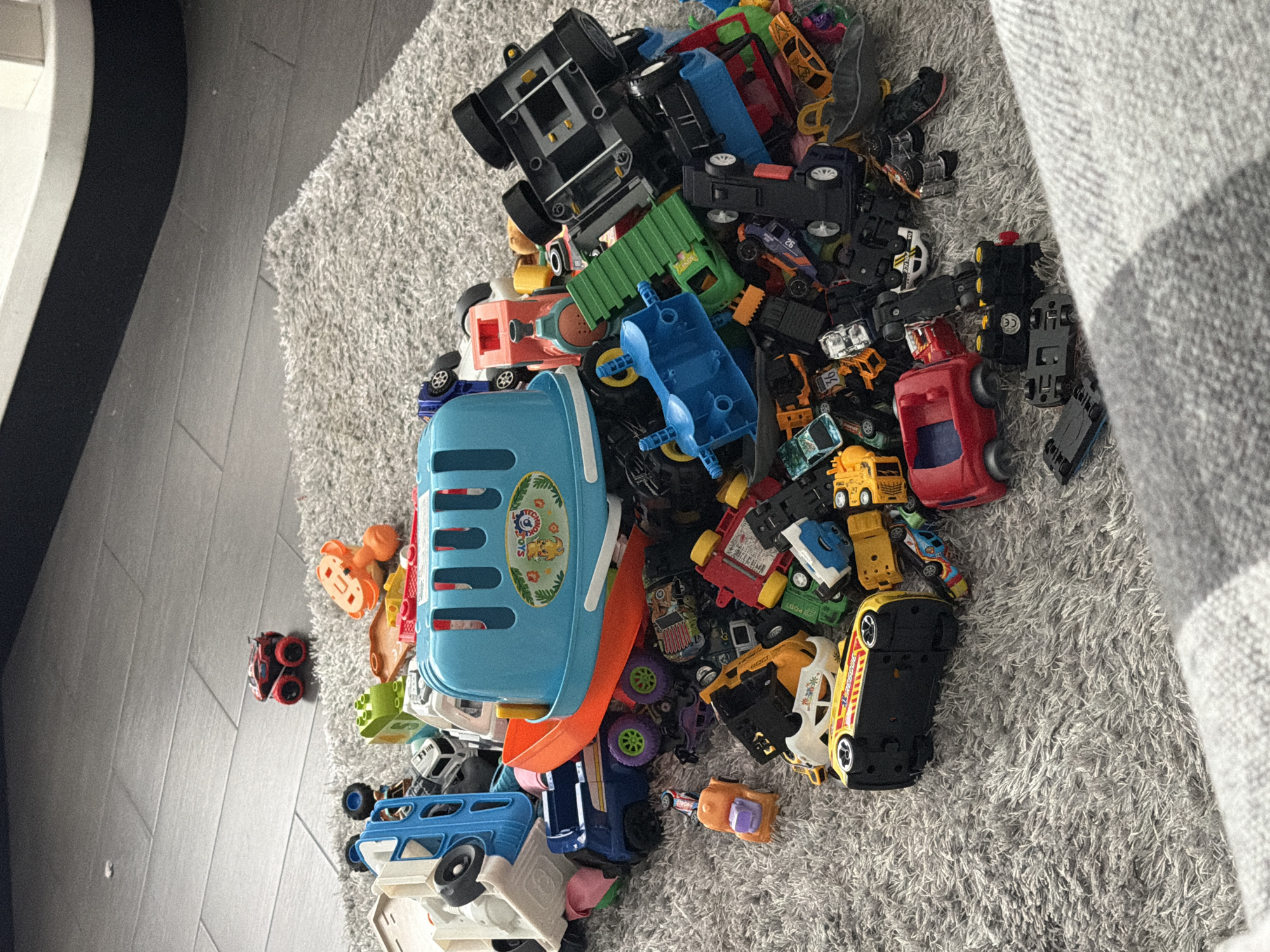 A pile of colorful toy vehicles is scattered across a gray, shag carpet. The toys include cars, trucks, and a blue plastic container that resembles a basket. The scene appears to be indoors, likely in a child's play area. The image does not contain any information that would allow for the identification of its location in Bila Tserkva, Ukraine, or any specific details about people, actions, or interactions.