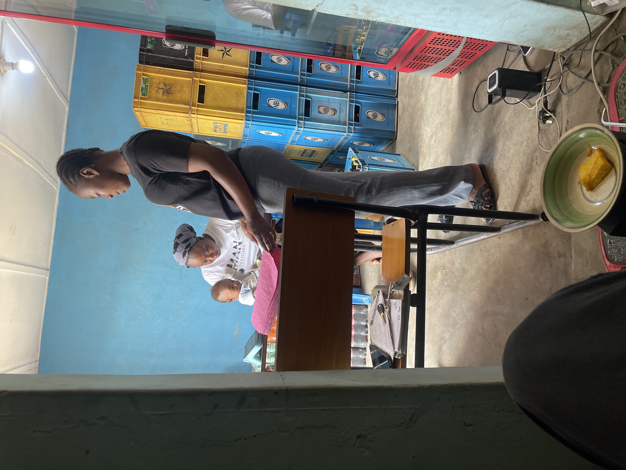 **Scene:** An indoor setting in Jos, Nigeria.
**People:** A young woman wearing a black t-shirt and grey pants leans over a table. Nearby, a woman wearing a white t-shirt and headscarf holds a baby.
**Objects:** Stacked beer crates in yellow and blue, a glass-fronted display case, a black power strip with multiple cables connected, a plate of food (possibly cake), a table, and a view of the outdoors from what appears to be a partially blocked opening.
**Environment:** The interior features blue walls and a concrete floor. Electrical cables are visible around the power strip.