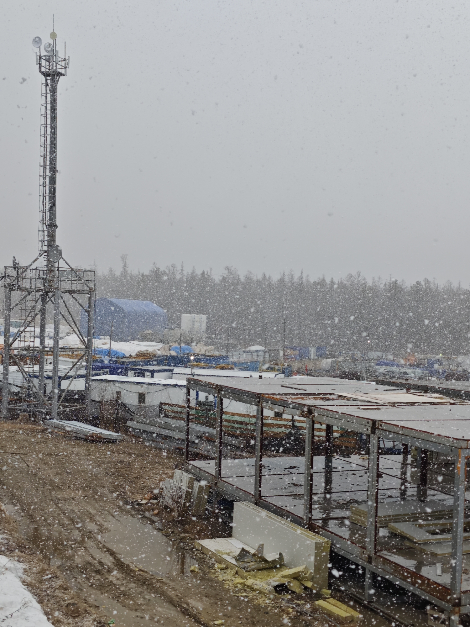 The image depicts a snowy, overcast day at what appears to be an industrial site or construction zone. A tall, metal communications tower stands prominently on the left side. In the background, a line of trees is visible. A partially constructed building or structure dominates the foreground, featuring a metal framework. Various building materials, including panels and insulation, are scattered around. The ground is wet and muddy, with patches of snow visible. There are no people visible in the scene. In the middle ground, there is an industrial-looking site containing a white and blue building tent among a pile of construction materials. The location is listed as being in the city of Unknown, Russia.